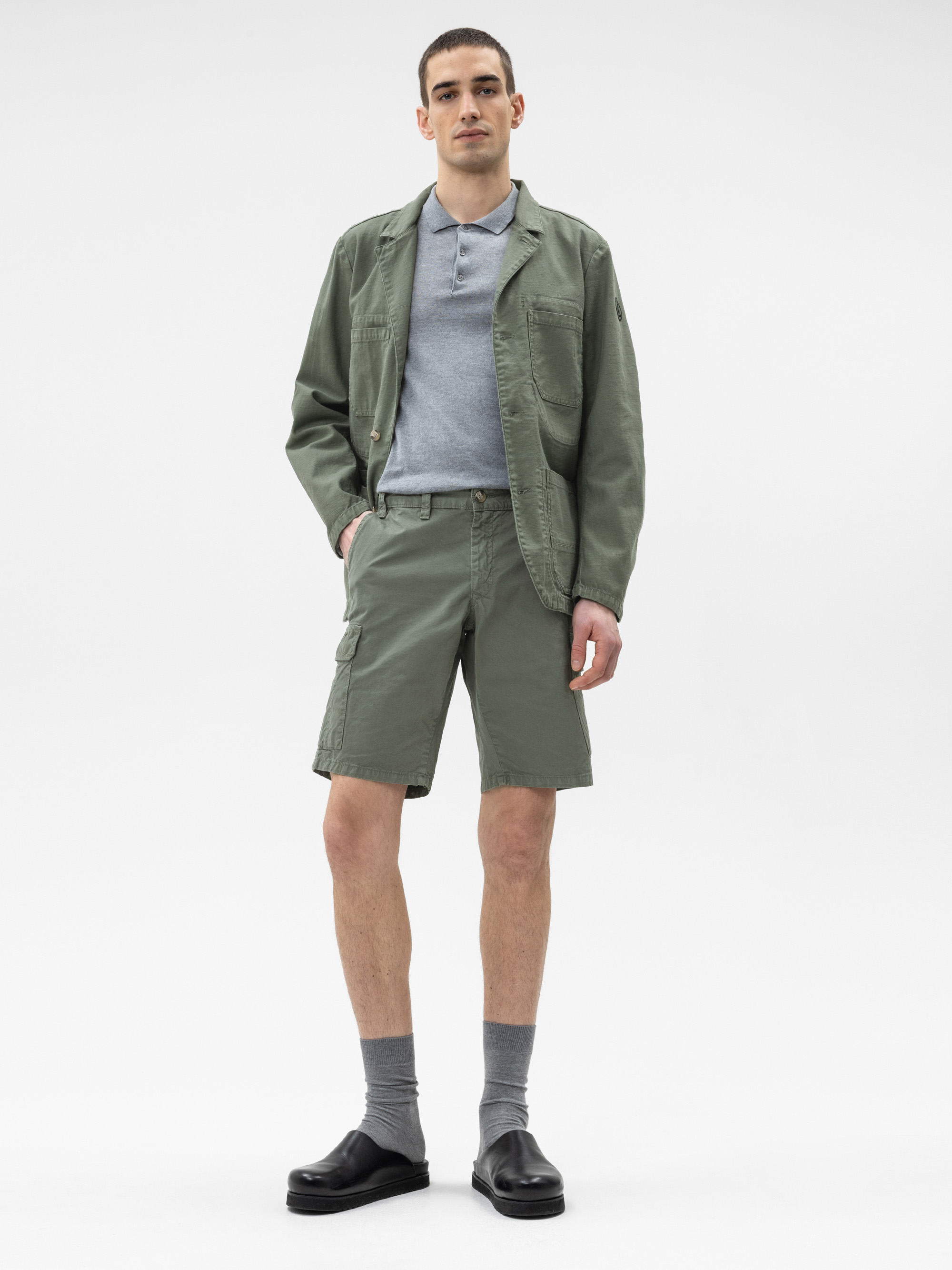 Ein Mann steht vor einem schlichten weißen Hintergrund und trägt eine salbeigrüne Jacke, passende Shorts, ein hellgraues Poloshirt, graue Socken und schwarze Slipper.