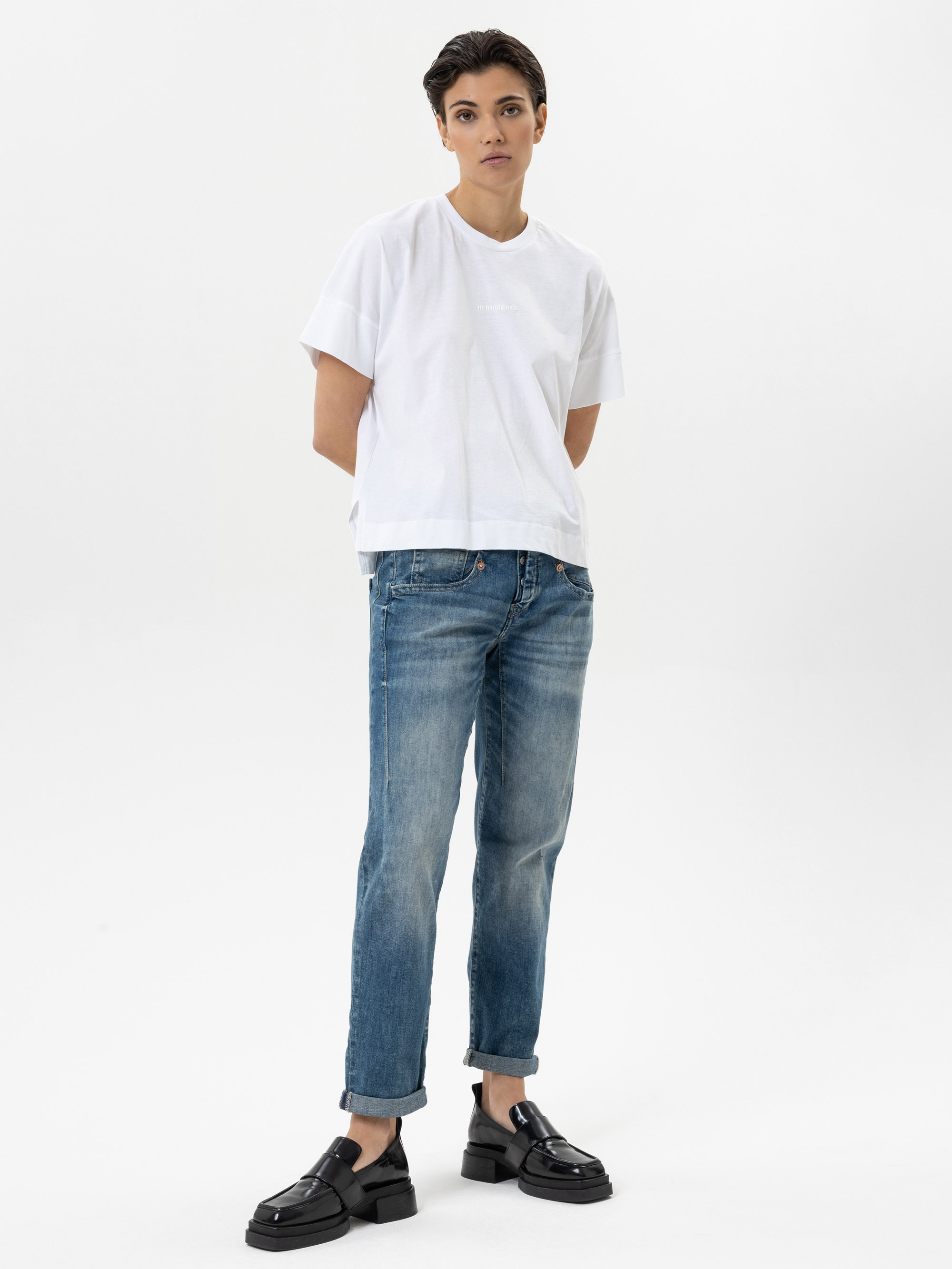 Eine Person mit kurzen dunklen Haaren, die ein lockeres weißes T-Shirt, blaue Jeans mit Manschetten und schwarze Slipper trägt, steht mit den Händen auf dem Rücken vor einem schlichten weißen Hintergrund.