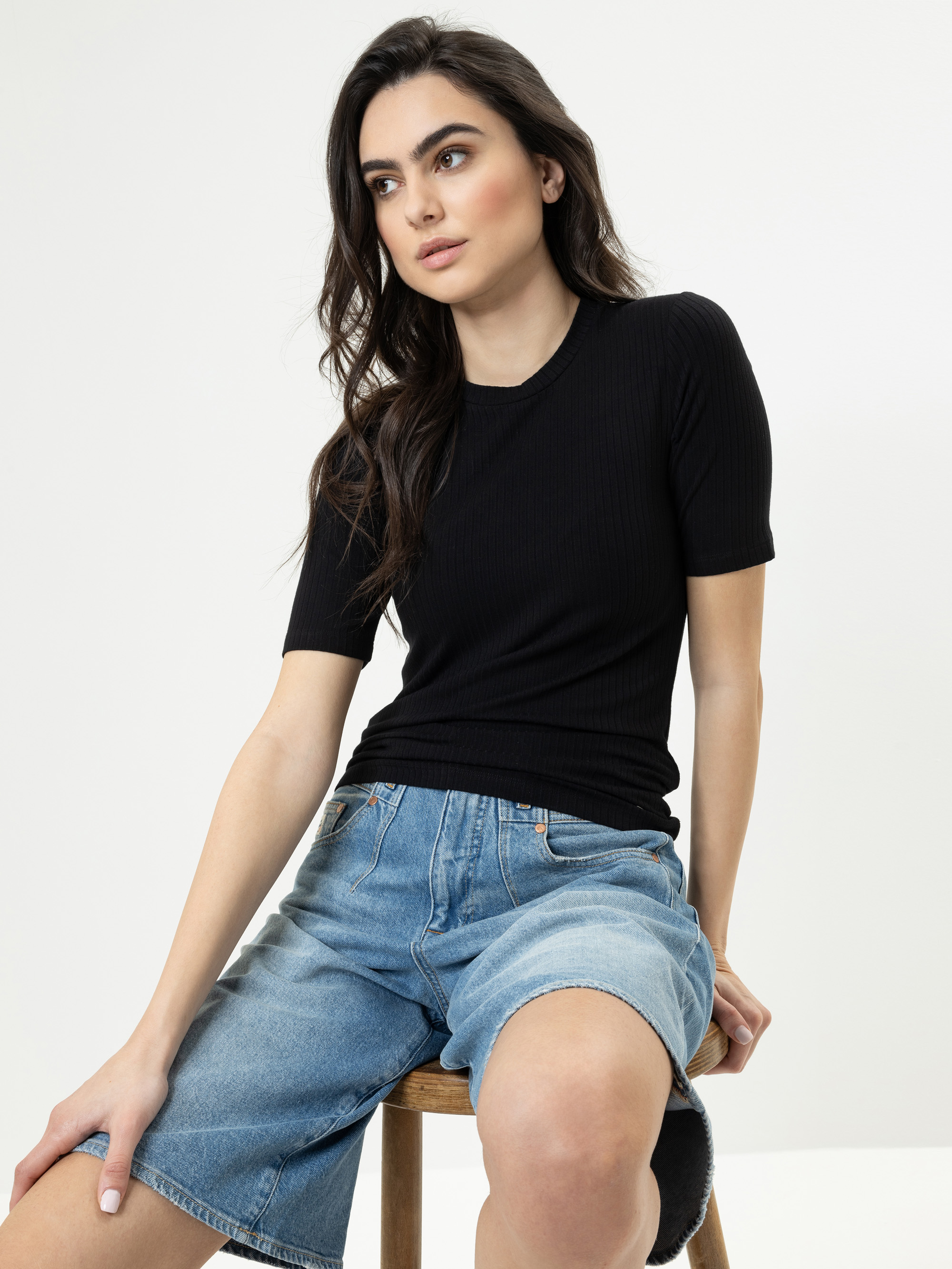 Eine Frau mit langen dunklen Haaren, die ein schwarzes kurzärmeliges Oberteil und blaue Jeansshorts trägt, sitzt auf einem Holzhocker vor einem schlichten weißen Hintergrund und schaut leicht zur Seite.