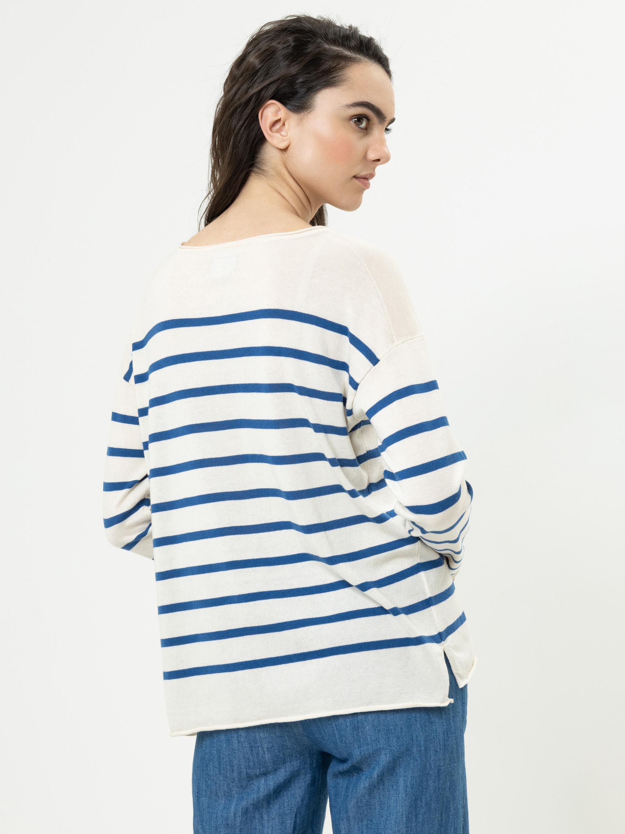 Eine Frau mit langen dunklen Haaren, die einen locker sitzenden, cremefarbenen Pullover mit blauen Querstreifen und eine blaue Jeans trägt, steht leicht von der Kamera abgewandt vor einem schlichten weißen Hintergrund.