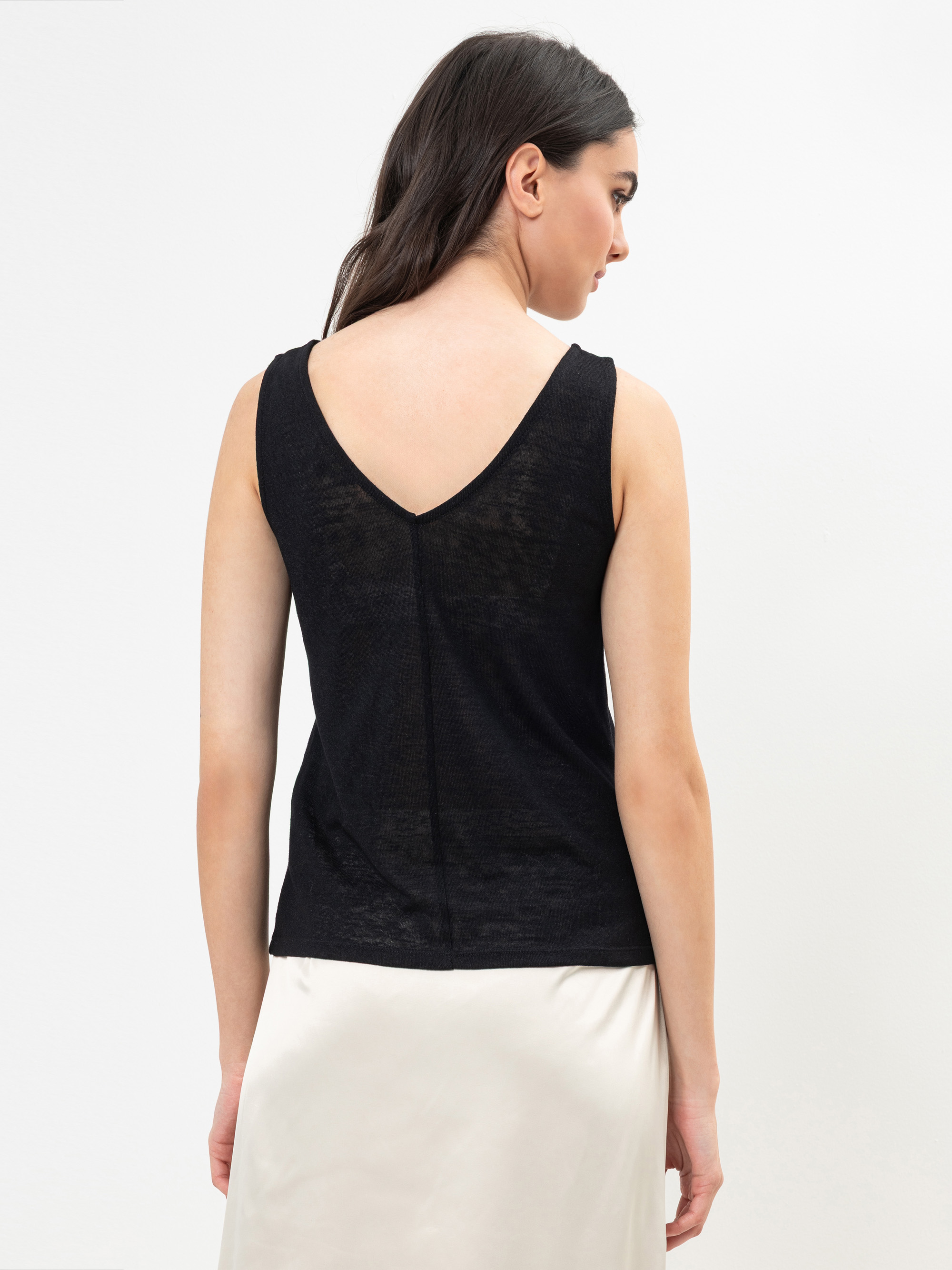Eine Frau mit langen dunklen Haaren trägt ein ärmelloses schwarzes Oberteil mit V-förmigem Rücken und einen hellen Rock. Sie steht vor einem schlichten weißen Hintergrund und ist von hinten fotografiert.