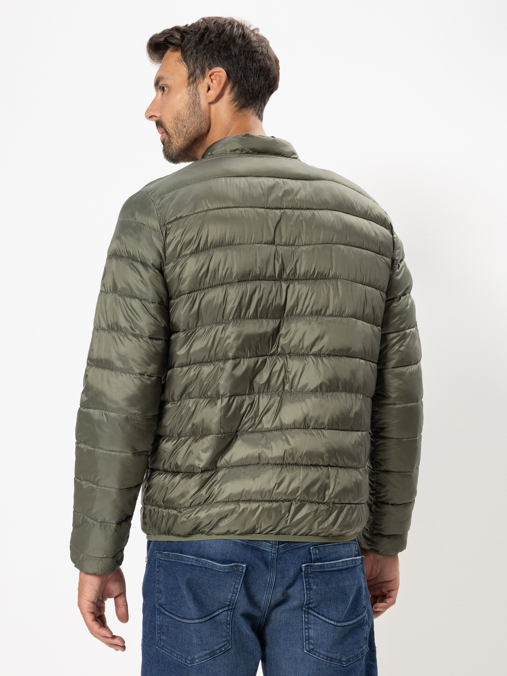 Ein Mann in grüner Steppjacke und blauer Jeans steht mit dem Gesicht zur Seite vor einem schlichten weißen Hintergrund.