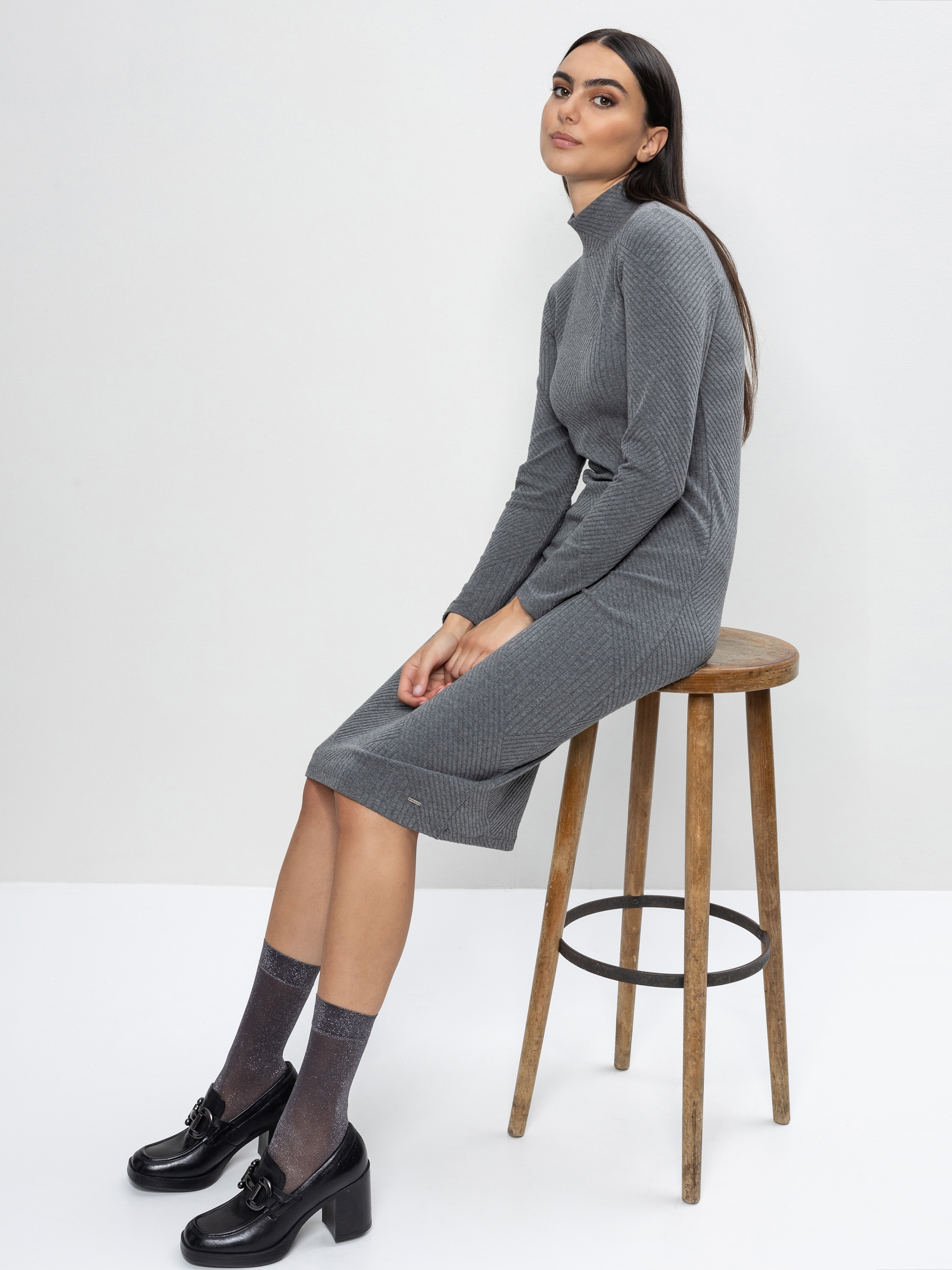 Eine Frau mit langen dunklen Haaren, die ein graues, geripptes Rollkragenkleid, glänzende schwarze Slipper und durchsichtige Socken trägt, sitzt seitlich auf einem Holzschemel vor einem schlichten hellgrauen Hintergrund.