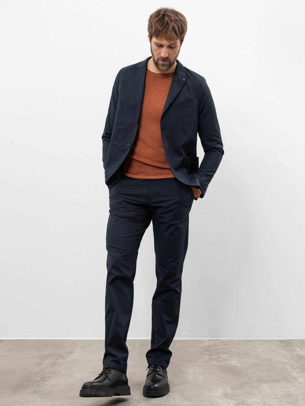 Ein Mann steht vor einer schlichten weißen Wand, trägt einen dunklen marineblauen Anzug mit einem rostfarbenen Pullover darunter und schwarze Anzugschuhe und schaut mit den Händen in den Taschen nach unten.