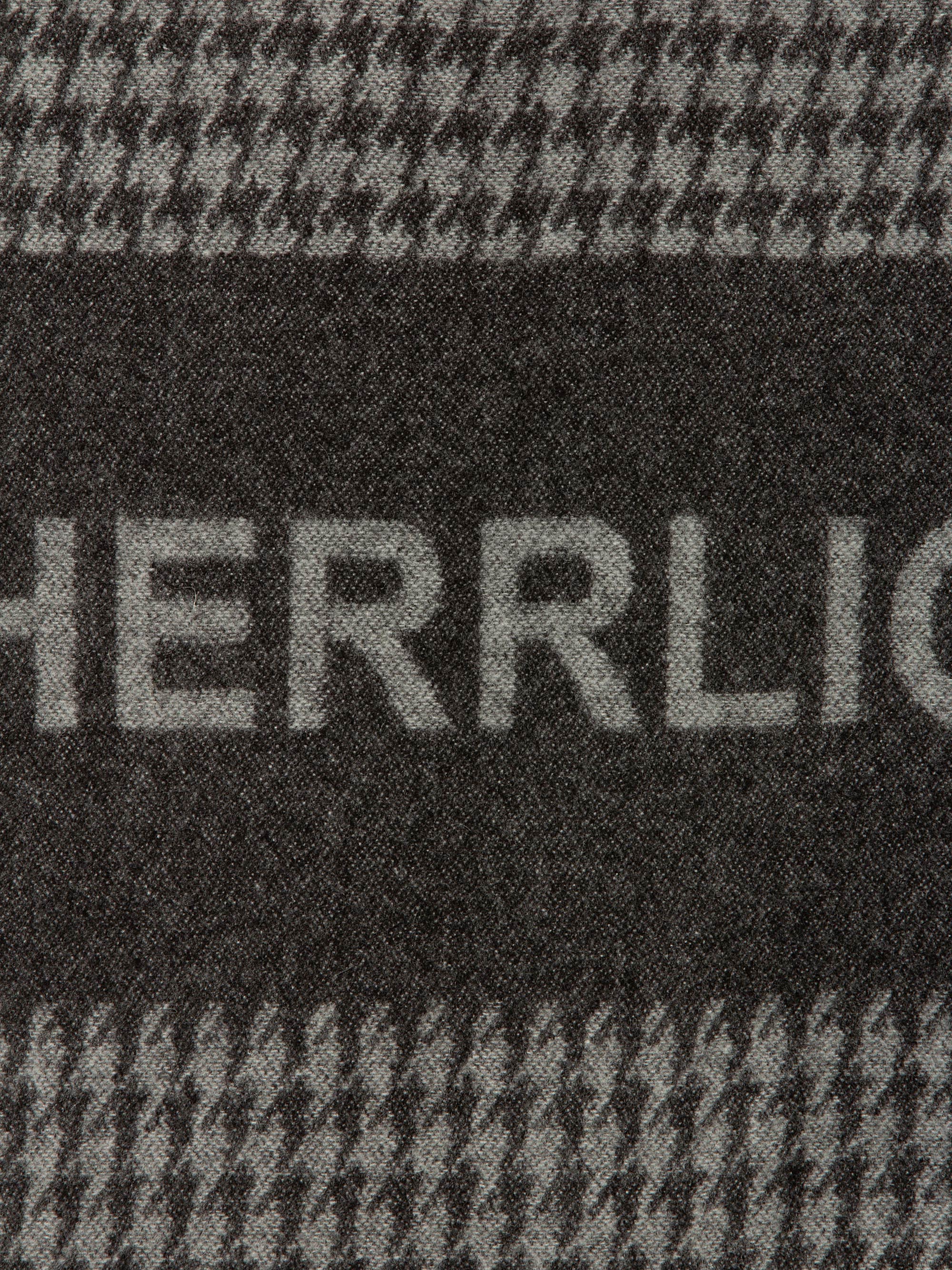 Nahaufnahme eines Stoffes mit Hahnentrittmuster in Schwarz und Grau, mit dem teilweise sichtbaren Wort "HERRLICH", das in hellerem Grau über einen dunklen Streifen gewebt ist.