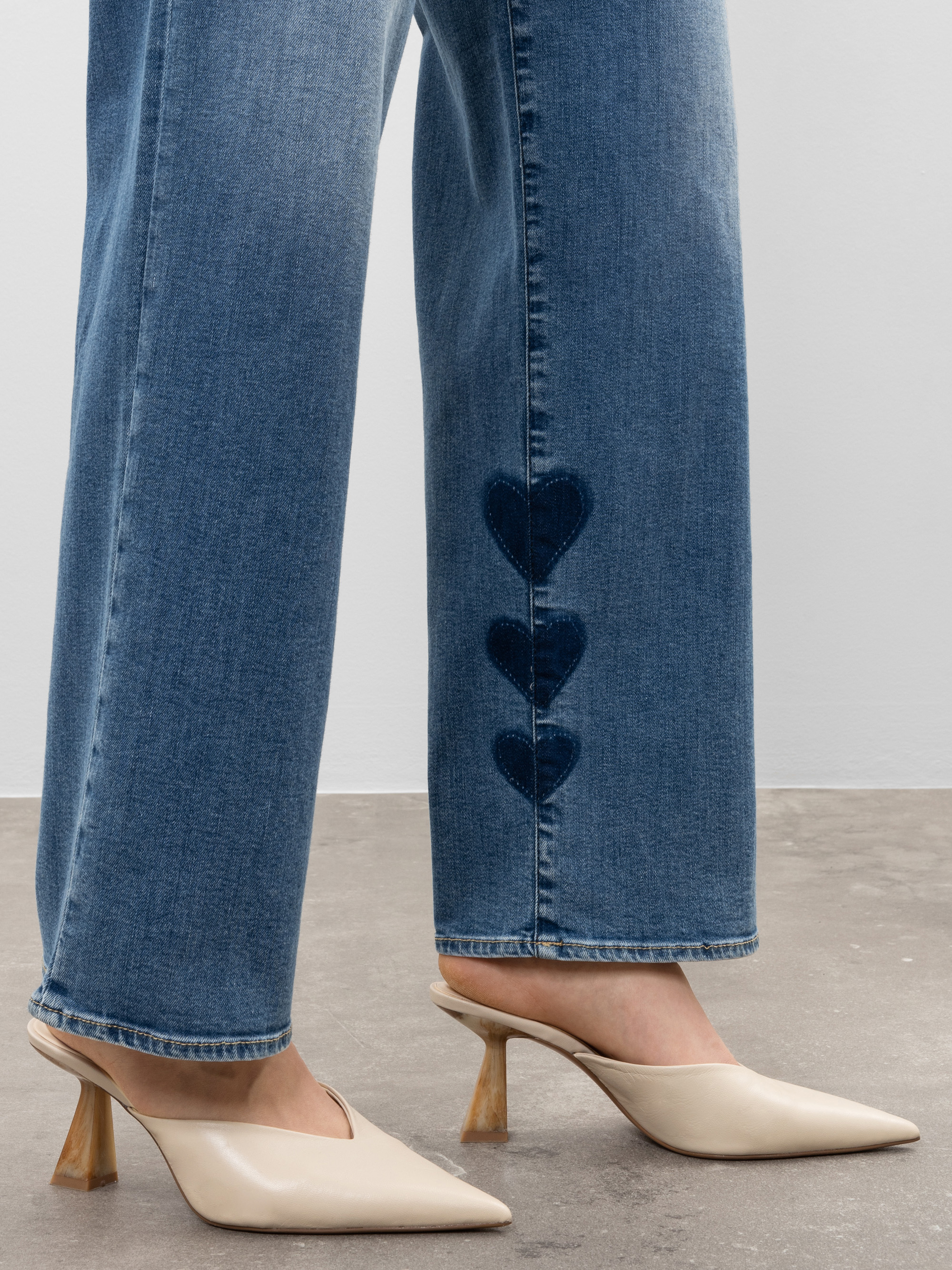 Eine Person, die eine hellblaue Jeans mit drei dunkelblauen herzförmigen Aufnähern an der Seite in der Nähe des Saums trägt, gepaart mit beigen Pantoletten mit spitzen Absätzen.