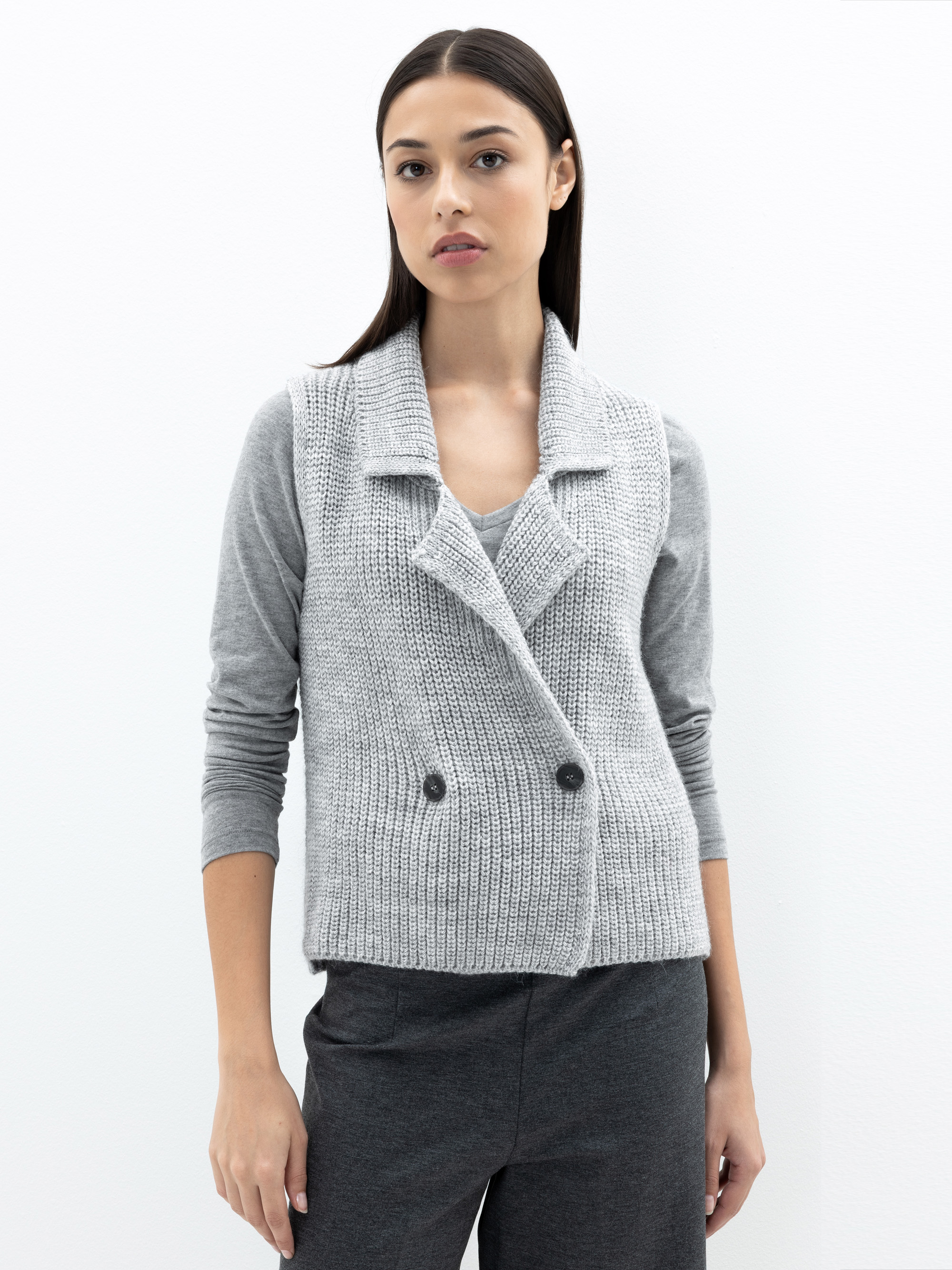 Eine Frau mit glattem dunklem Haar trägt eine hellgraue Strickweste über einem grauen langärmeligen Oberteil und eine dazu passende graue Hose vor einem schlichten weißen Hintergrund.