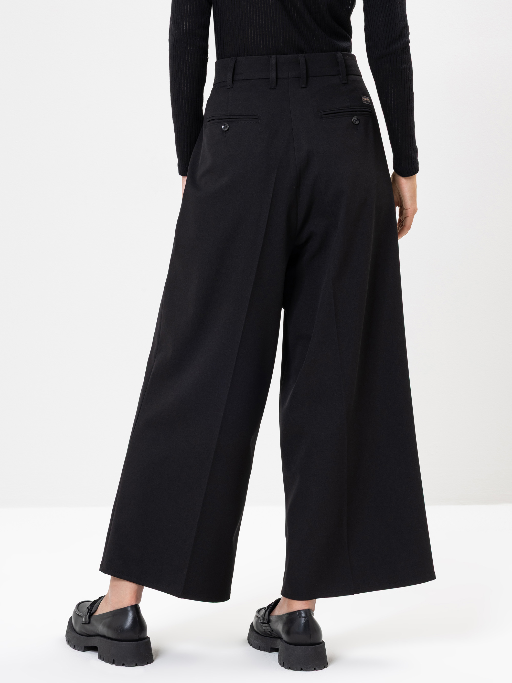 Eine Person, die eine schwarze Hose mit weitem Bein, ein schwarzes, langärmeliges Oberteil und schwarze, klobige Schuhe trägt, steht mit dem Gesicht nach unten auf einem weißen Hintergrund.