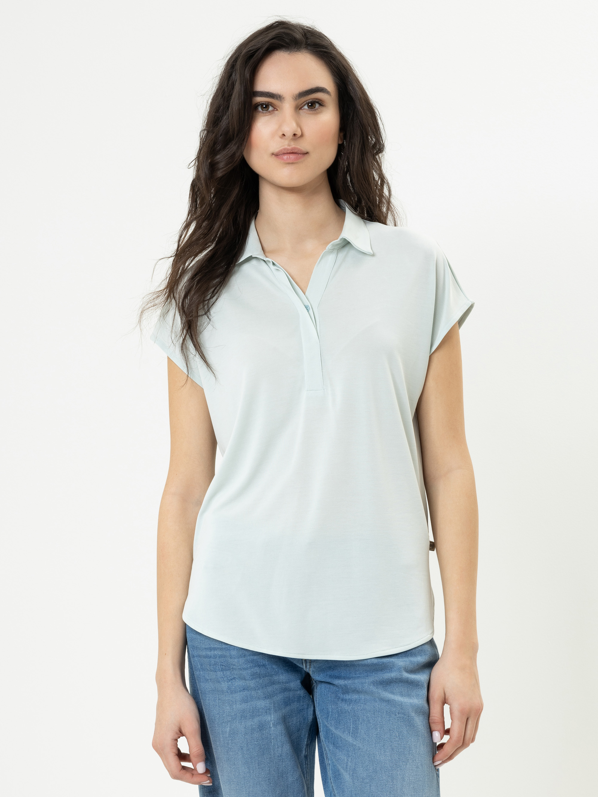 Eine Frau mit langen, gewellten dunklen Haaren trägt ein hellblaues kurzärmeliges Hemd mit Kragen und blaue Jeans. Sie steht vor einem schlichten weißen Hintergrund und hat einen neutralen Gesichtsausdruck.