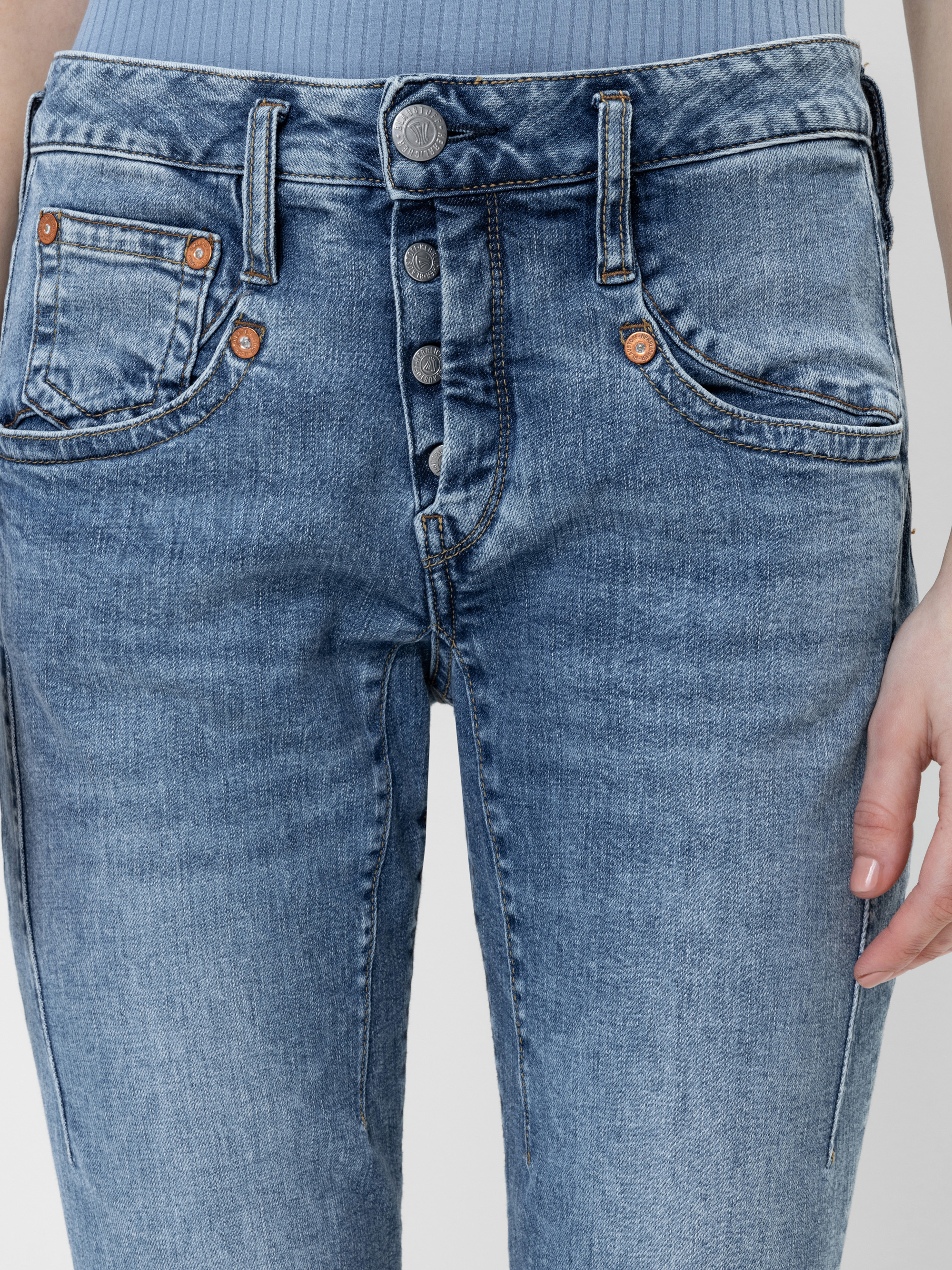 Nahaufnahme einer Person, die eine hellblaue Jeans mit hoher Taille, Knopfleiste und kupferfarbenen Nieten trägt, dazu ein hellblaues, geripptes Oberteil zum Hineinstecken. Nur der untere Teil des Oberkörpers und eine Hand sind zu sehen.