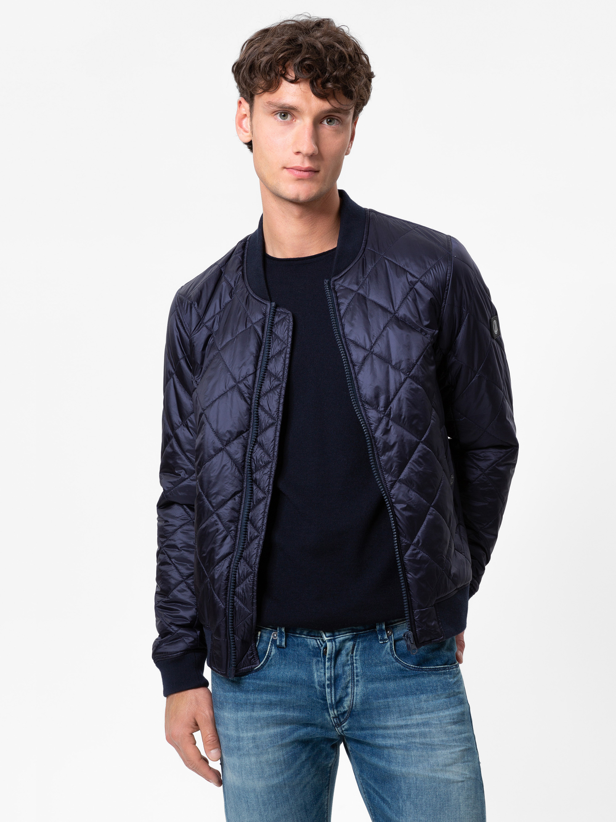Ein junger Mann mit lockigem braunem Haar trägt eine marineblaue gesteppte Bomberjacke über einem schwarzen Hemd und blauen Jeans und steht vor einem schlichten weißen Hintergrund.