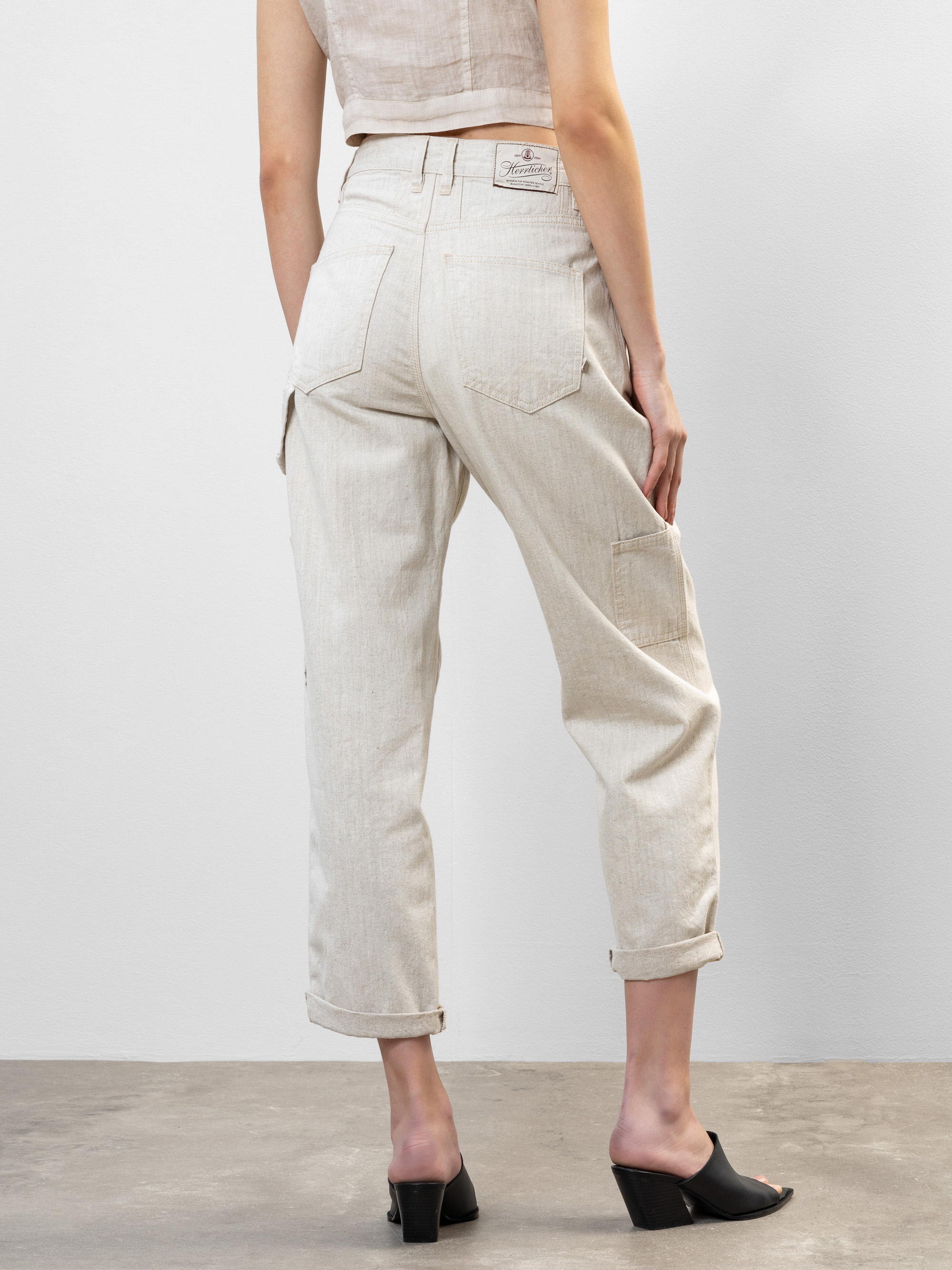 Eine Person steht mit dem Gesicht nach hinten und trägt eine hellbeige Hose mit Bündchen und Gesäßtaschen sowie ein ärmelloses Oberteil. Außerdem trägt sie schwarze, zehenoffene Sandalen mit Absatz. Der Hintergrund ist schlicht und minimalistisch.