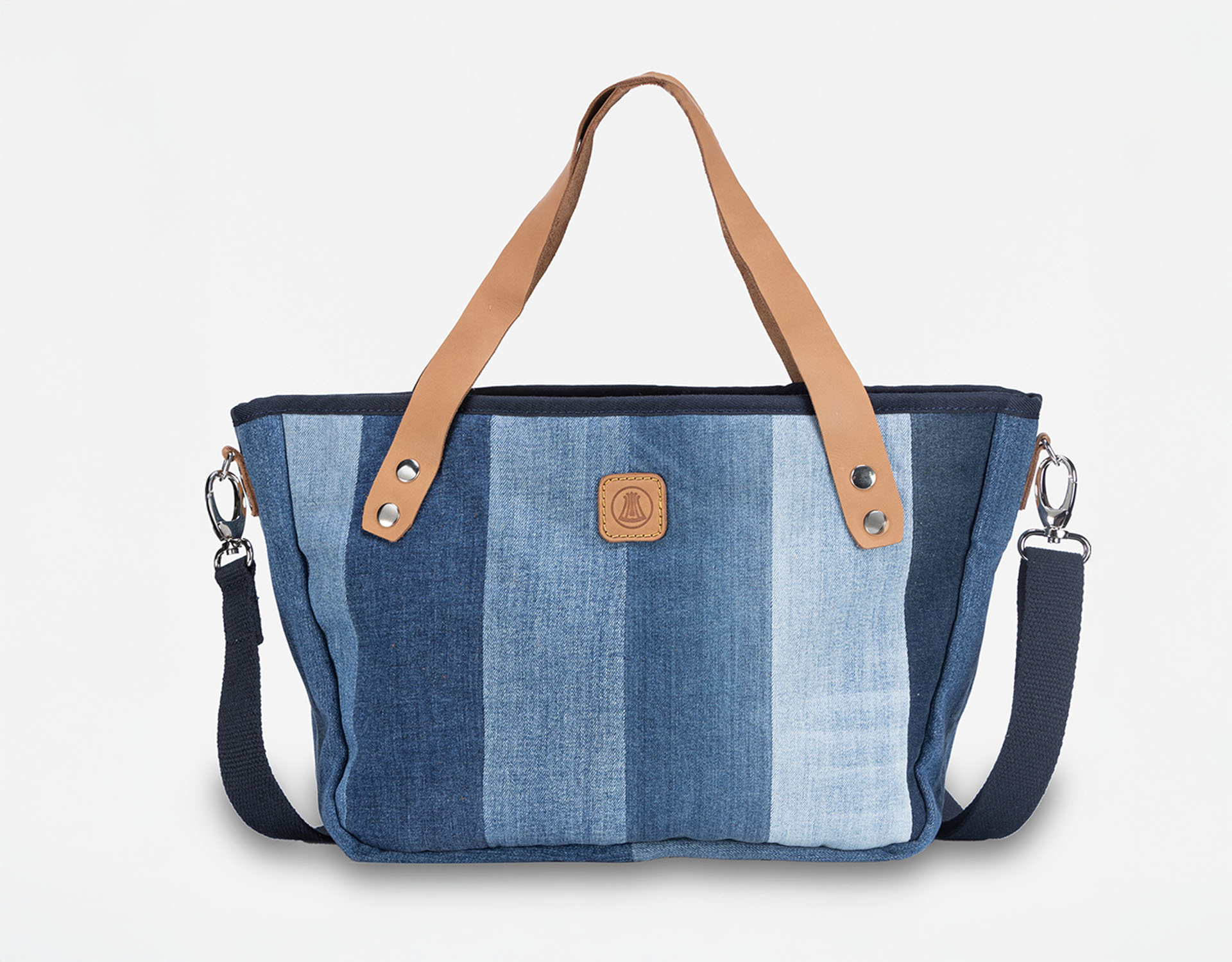 Eine stilvolle Tragetasche mit vertikalen blauen Denim-Einsätzen, hellbraunen Ledergriffen, einem abnehmbaren dunkelblauen Schulterriemen und einem kleinen braunen Logoaufnäher auf der Vorderseite vor einem schlichten weißen Hintergrund.