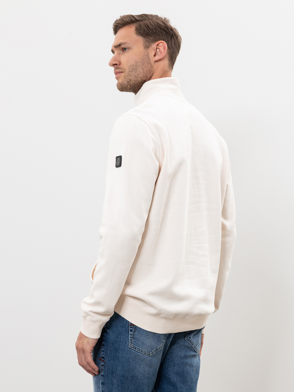 Ein Mann mit kurzen braunen Haaren trägt eine cremefarbene Jacke mit Reißverschluss und blaue Jeans. Er steht seitlich und blickt über seine Schulter vor einem schlichten weißen Hintergrund.