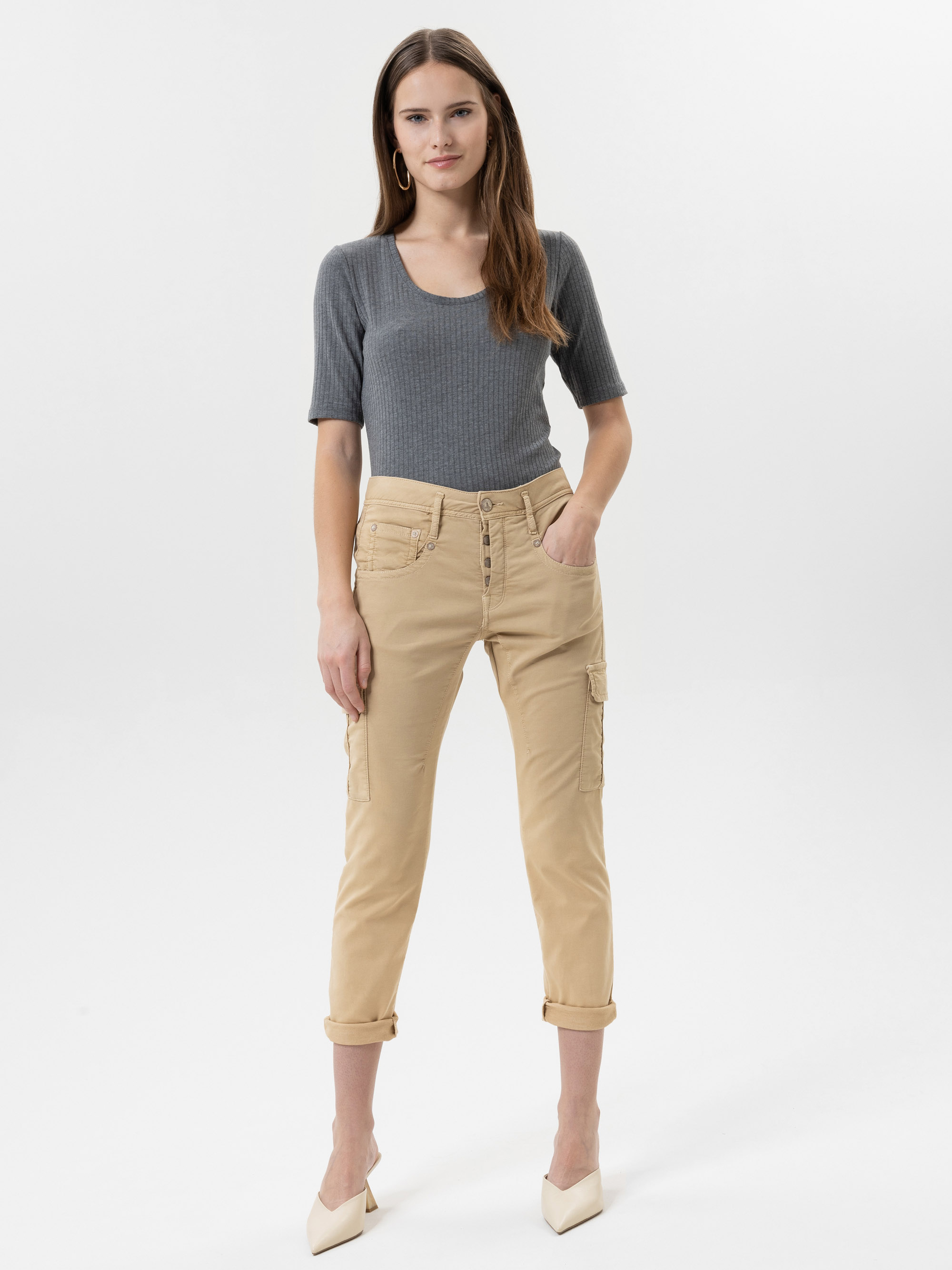 Eine Frau steht vor einem schlichten weißen Hintergrund und trägt ein graues, kurzärmeliges, geripptes Oberteil, eine beigefarbene, kurze Cargohose und weiße, spitze Absätze. Sie hat langes braunes Haar und eine entspannte, selbstbewusste Körperhalt