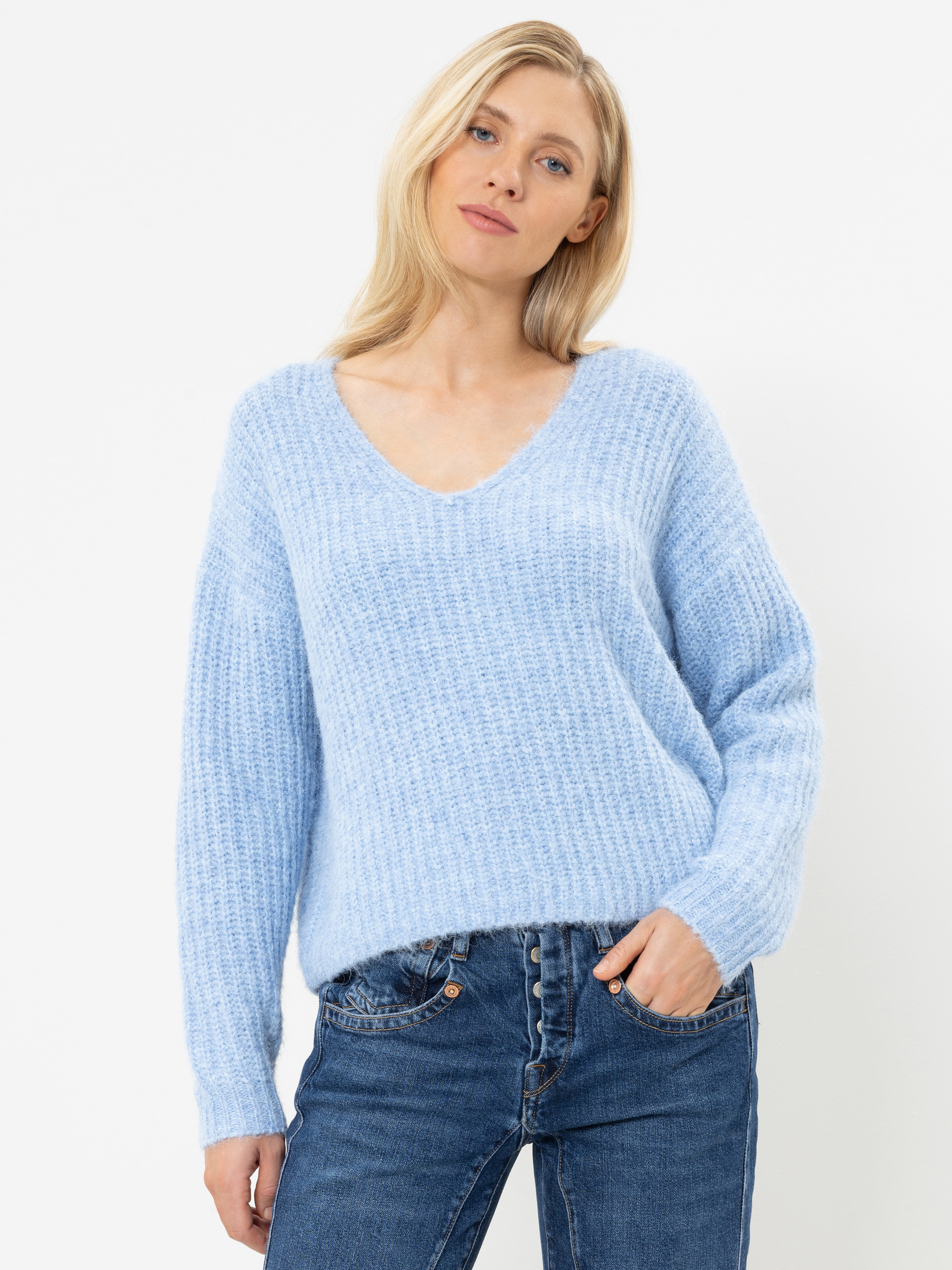Eine Frau mit blonden Haaren, die einen hellblauen, strukturierten Pullover mit V-Ausschnitt und eine blaue Jeans trägt, steht vor einem schlichten weißen Hintergrund, eine Hand in der Tasche und mit entspanntem Gesichtsausdruck.