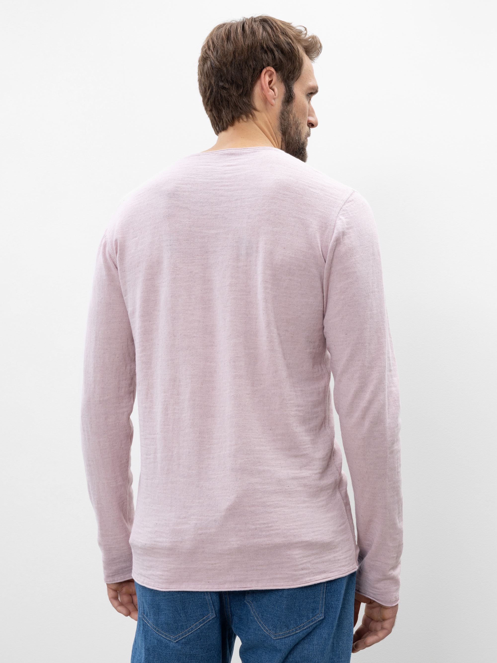 Ein Mann mit braunen Haaren, von hinten gesehen, trägt ein hellrosa langärmeliges Hemd und blaue Jeans vor einem schlichten weißen Hintergrund.