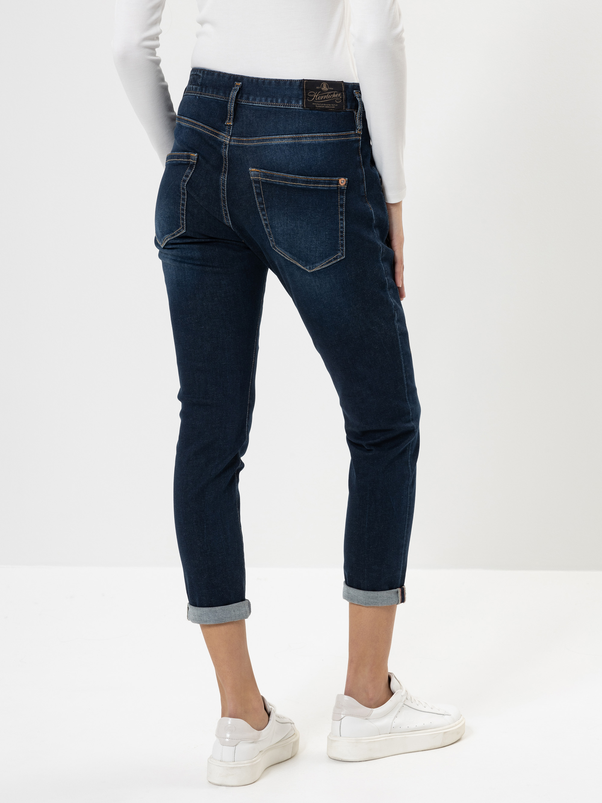 Eine Person, die dunkelblaue Jeans mit Bündchen, ein weißes langärmeliges Oberteil und weiße Turnschuhe trägt, steht mit dem Gesicht zur Kamera vor einem einfarbig weißen Hintergrund.