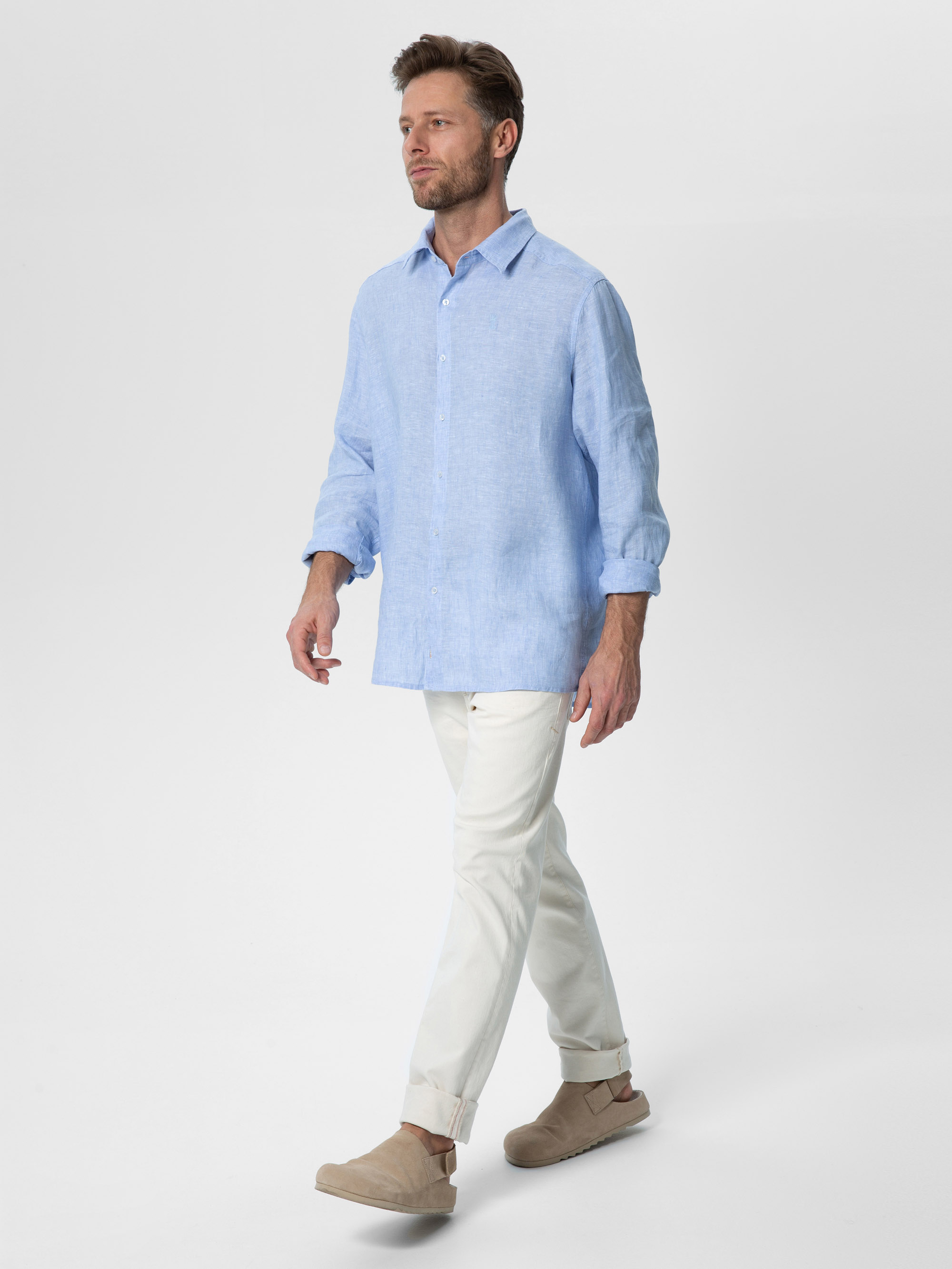 Ein Mann geht selbstbewusst in einem Studio spazieren. Er trägt ein hellblaues, langärmeliges Hemd, weiße, hochgekrempelte Hosen und beigefarbene Slipper. Der Hintergrund ist einfarbig weiß.