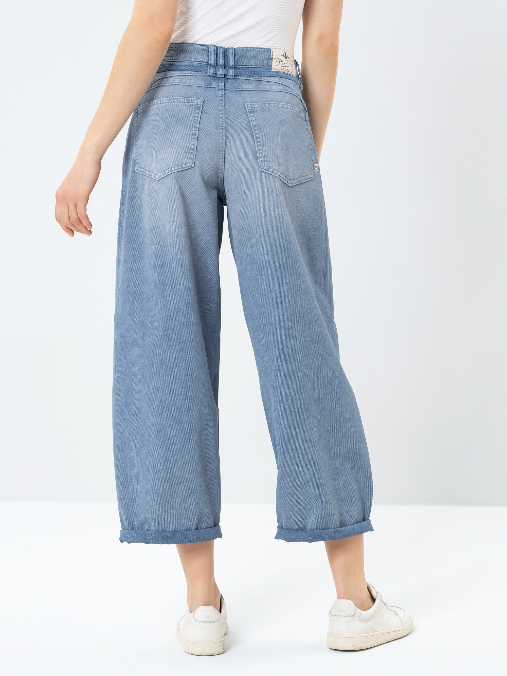 Eine Person, die hellblaue, weit geschnittene Jeans mit Gürtel und weißen Turnschuhen trägt, wird von hinten vor einem einfarbig weißen Hintergrund gezeigt.