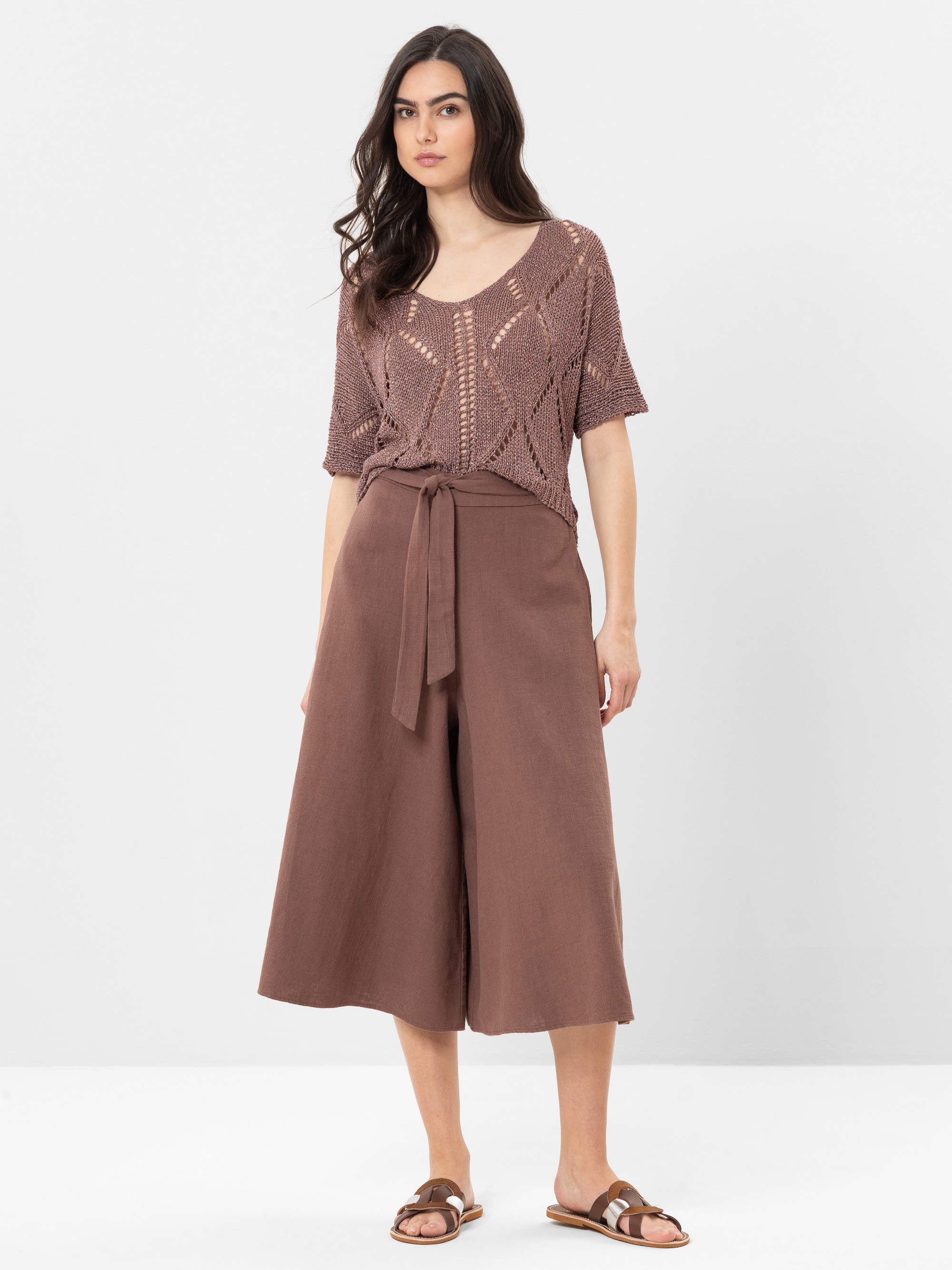 Eine Frau steht vor einem unifarbenen Hintergrund und trägt ein taupefarbenes, kurzärmeliges Strickoberteil, eine weit geschnittene braune Culotte-Hose mit Bindeband und braune Sandalen. Sie hat langes dunkles Haar und einen neutralen Gesichtsausdruck.