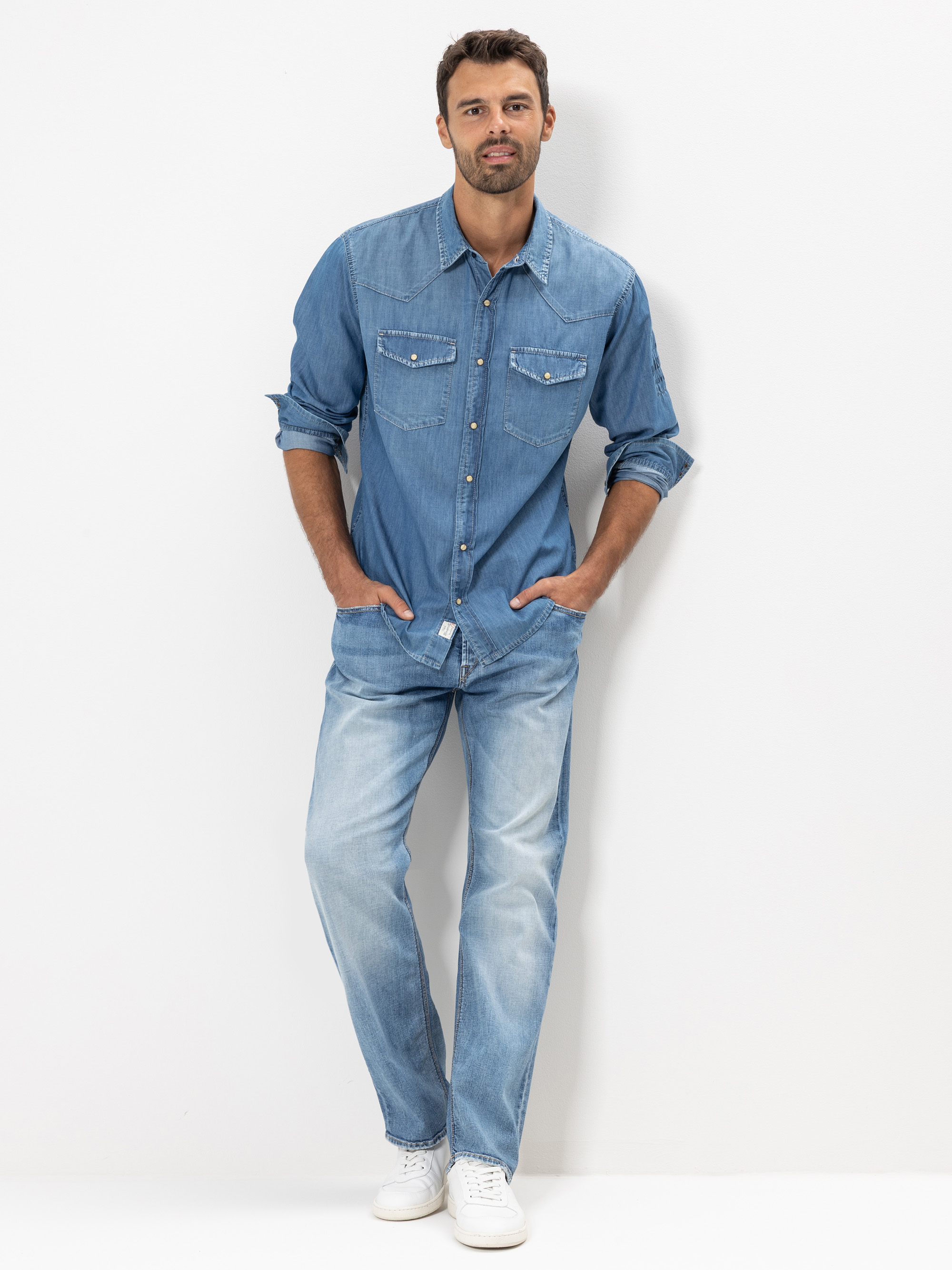 Ein Mann steht vor einem weißen Hintergrund und trägt ein hellblaues Jeanshemd, hellblaue Jeans und weiße Turnschuhe. Seine Hände stecken in den Taschen und er lächelt leicht.