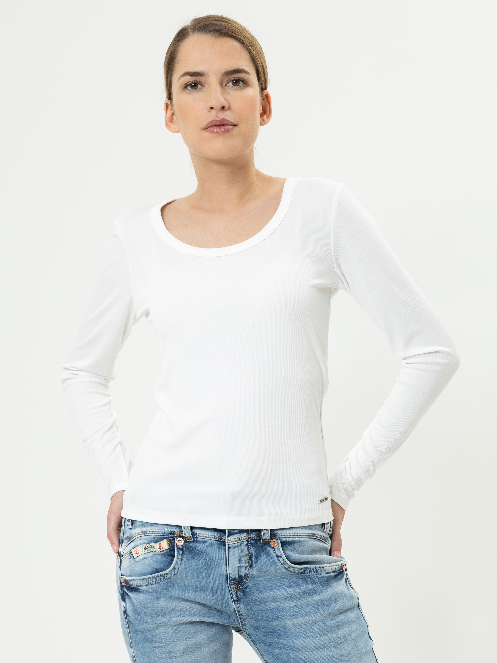 Eine Frau in einem taillierten, langärmeligen weißen Oberteil und einer blauen Jeans steht vor einem schlichten weißen Hintergrund, hat die Hände in den Taschen und blickt selbstbewusst nach vorn.