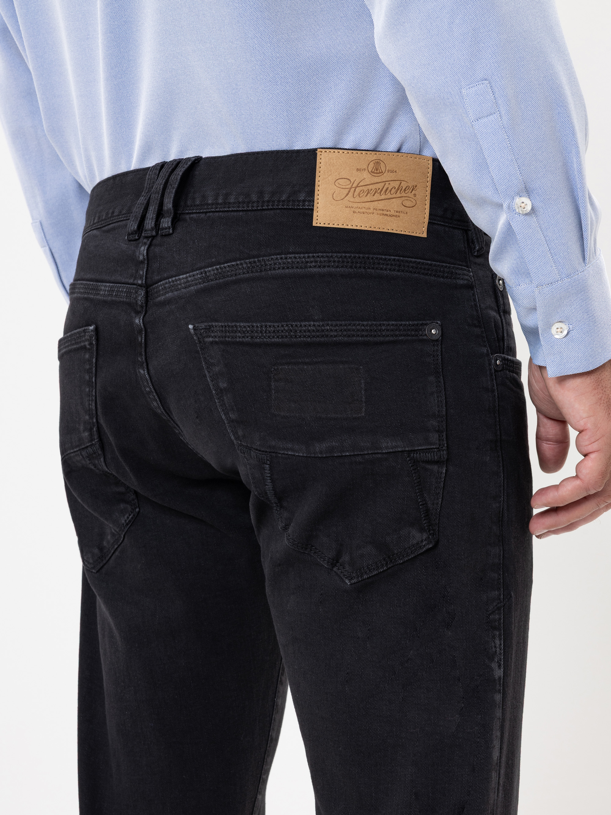 Eine Person trägt eine schwarze Jeans und ein hellblaues Hemd, von hinten gesehen. Die Jeans hat ein braunes Lederetikett am Bund und verfügt über zwei Gesäßtaschen mit dezenten Nähten.