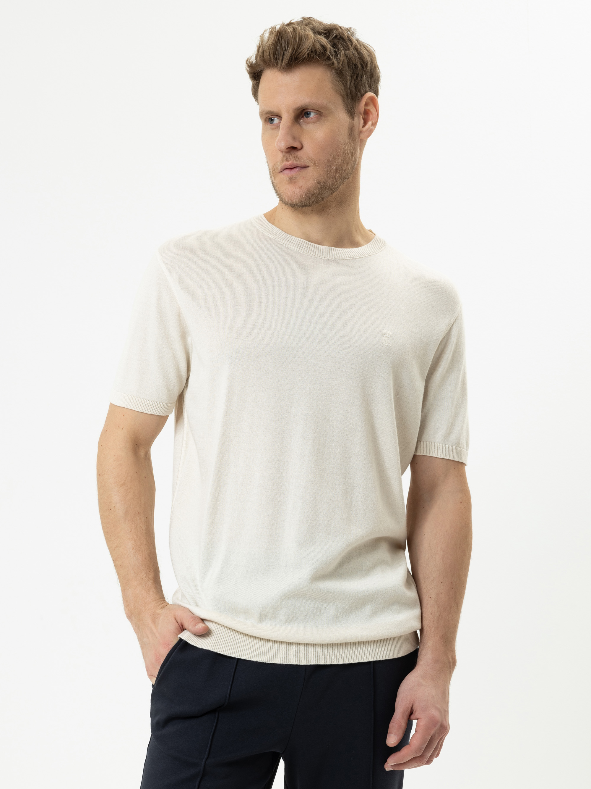 Ein Mann mit kurzen, hellbraunen Haaren trägt einen hellbeigen, kurzärmeligen Strickpullover und eine dunkle Hose und steht mit einer Hand in der Tasche vor einem schlichten weißen Hintergrund.