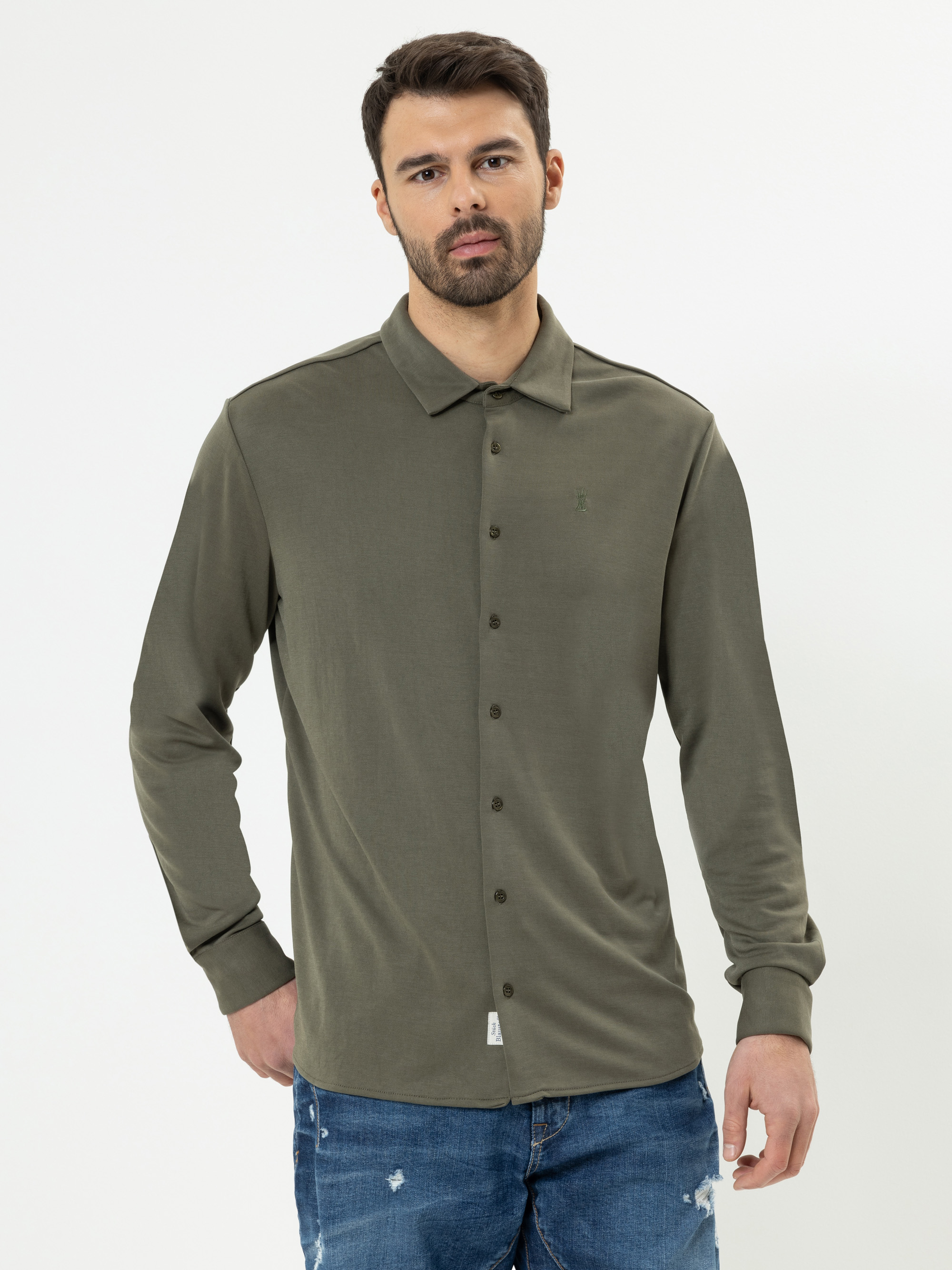 Ein Mann mit kurzen dunklen Haaren und einem Bart trägt ein olivgrünes Button-up-Hemd und blaue Jeans und steht vor einem schlichten weißen Hintergrund.