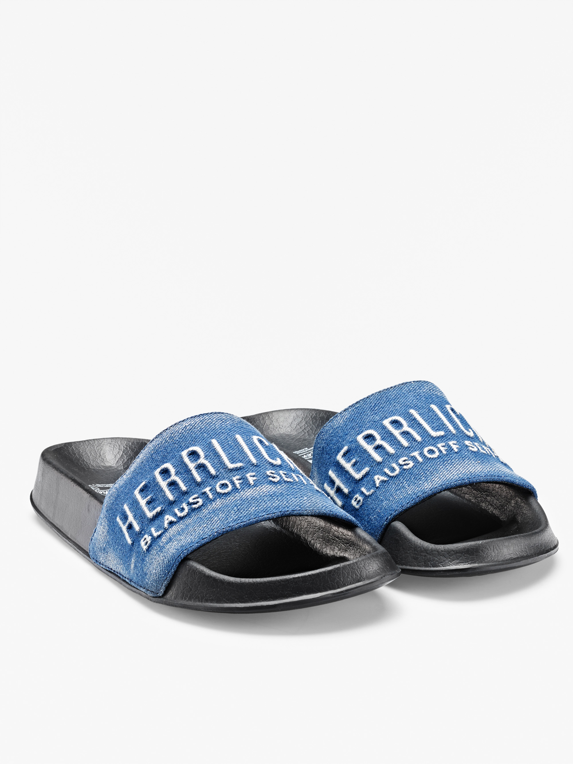 Ein Paar schwarze Gleitsandalen mit blauen Jeansriemen. Auf den Riemen ist der Schriftzug HERRLICH BLAUSTOFF SEIT weiß aufgestickt. Der Hintergrund ist einfarbig weiß.
