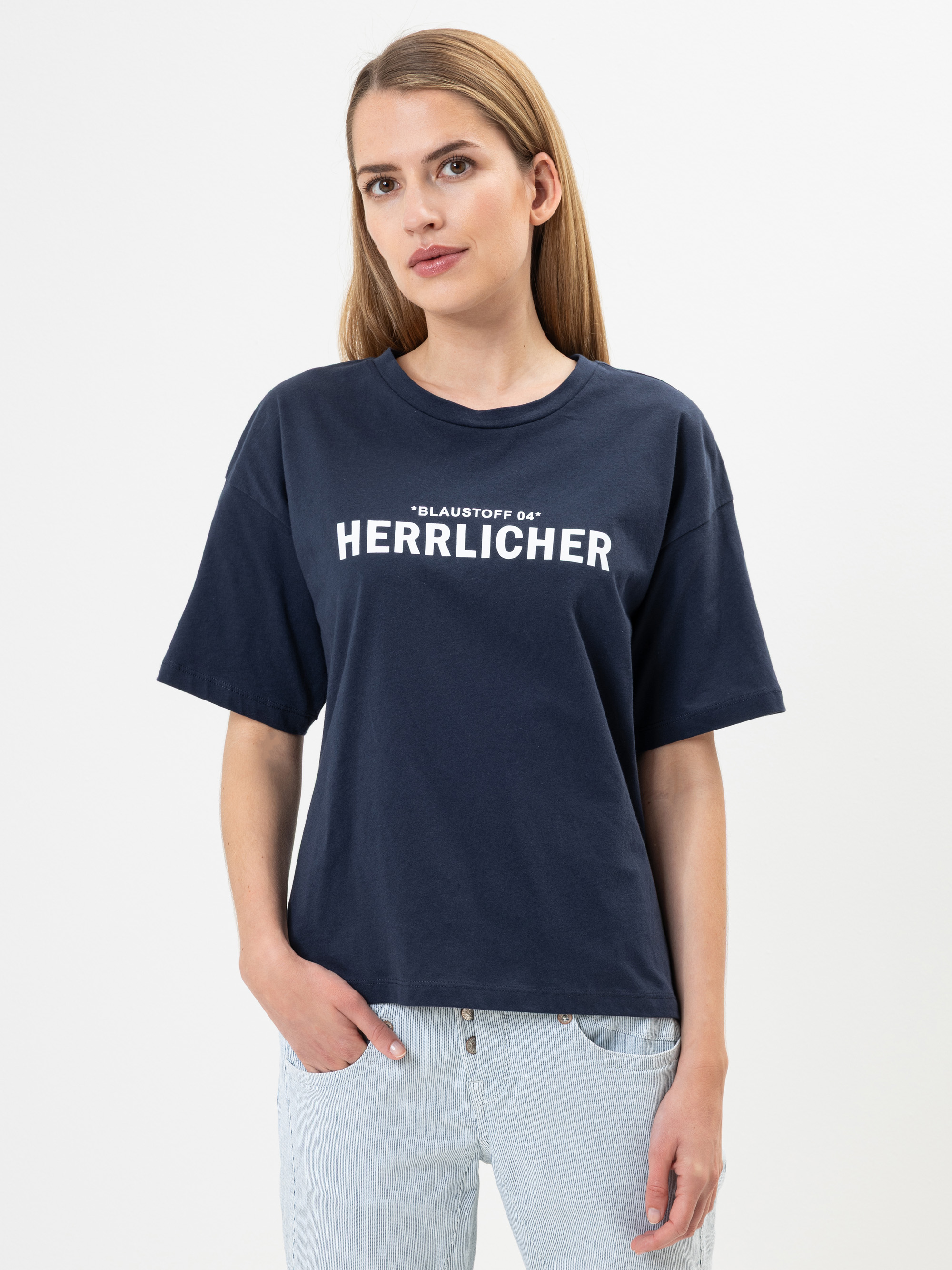 Eine Frau mit langem glattem Haar trägt ein marineblaues T-Shirt mit der Aufschrift BLAUSTOFF 04 HERRLICHER und hellblaue Jeans. Sie steht vor einem schlichten weißen Hintergrund und hat eine Hand in der Tasche.