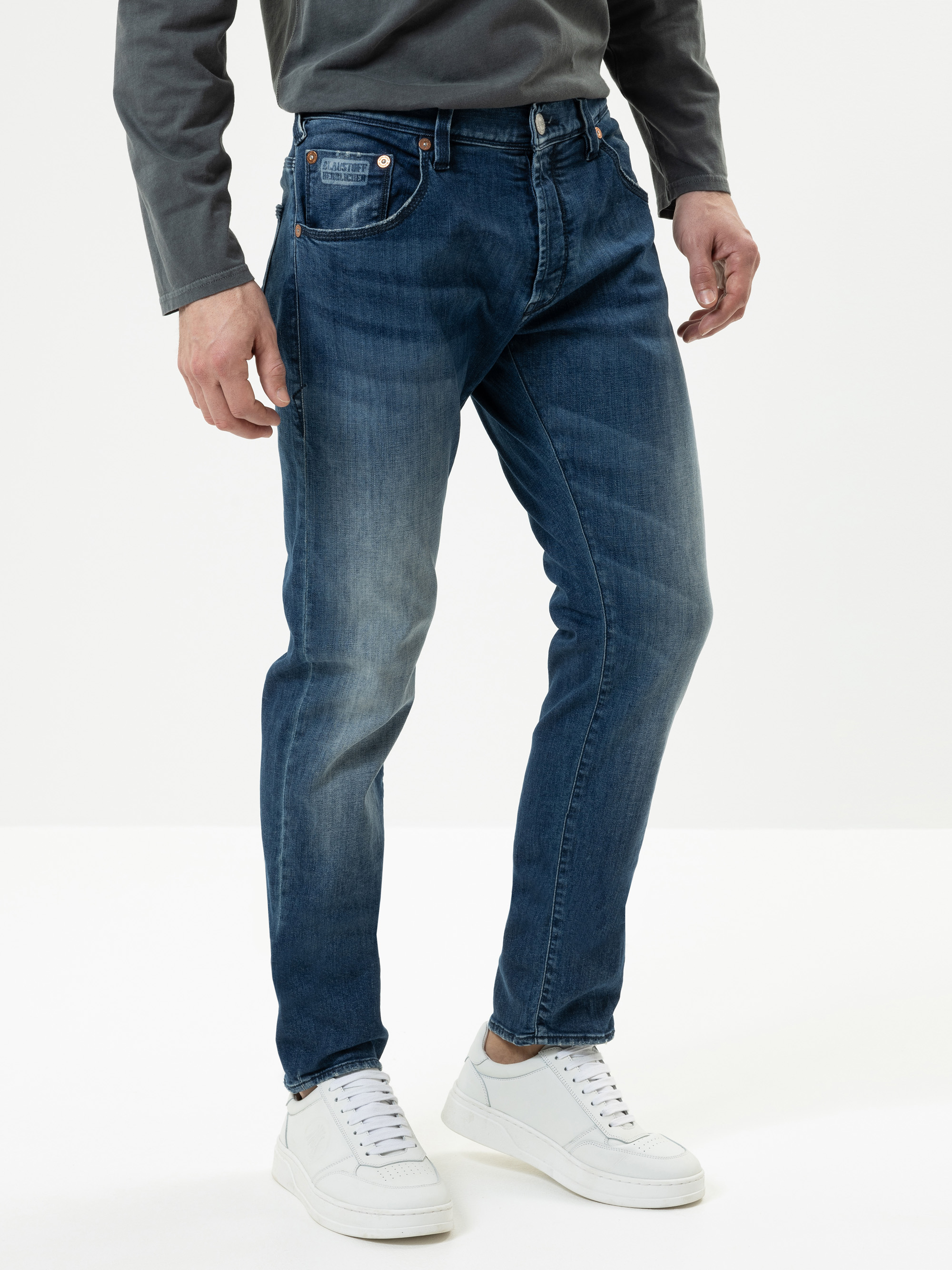 Eine Person, die dunkelblaue Jeans, ein graues langärmeliges Hemd und weiße Turnschuhe trägt, steht vor einem schlichten weißen Hintergrund. Nur der Unterkörper ist sichtbar.