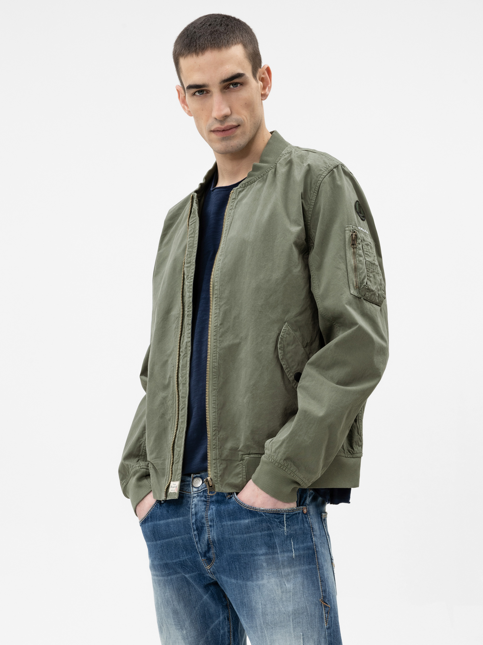 Ein Mann, der eine hellolivgrüne Bomberjacke über einem dunklen Hemd und eine blaue Jeans trägt, steht mit den Händen in den Taschen und blickt in die Kamera vor einem schlichten weißen Hintergrund.