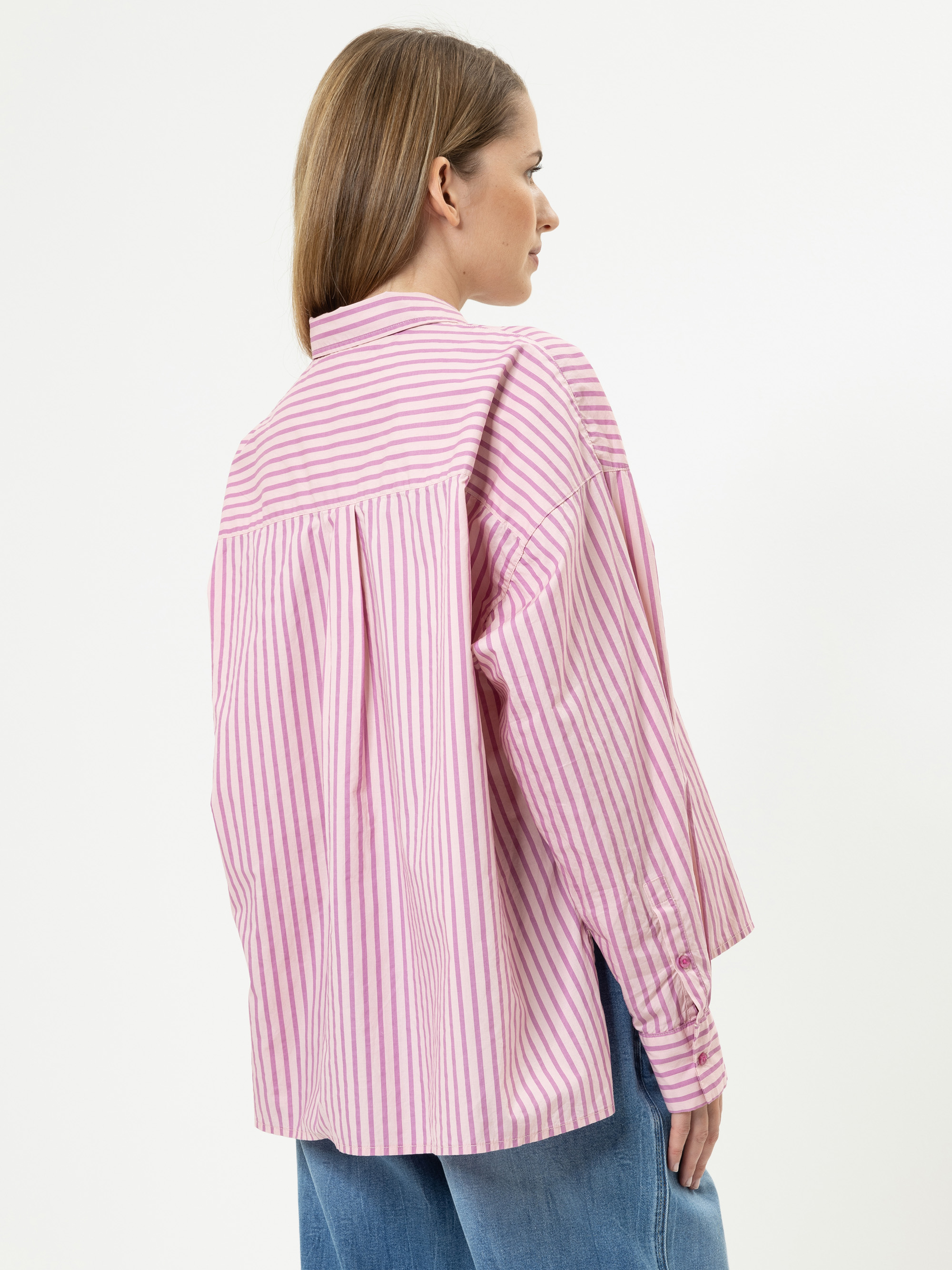 Eine Frau mit glattem, hellbraunem Haar trägt ein locker sitzendes, langärmeliges, rosa-weiß gestreiftes Hemd und blaue Jeans. Sie steht leicht von der Kamera abgewandt, so dass der Rücken und die Seite des Shirts zu sehen sind.