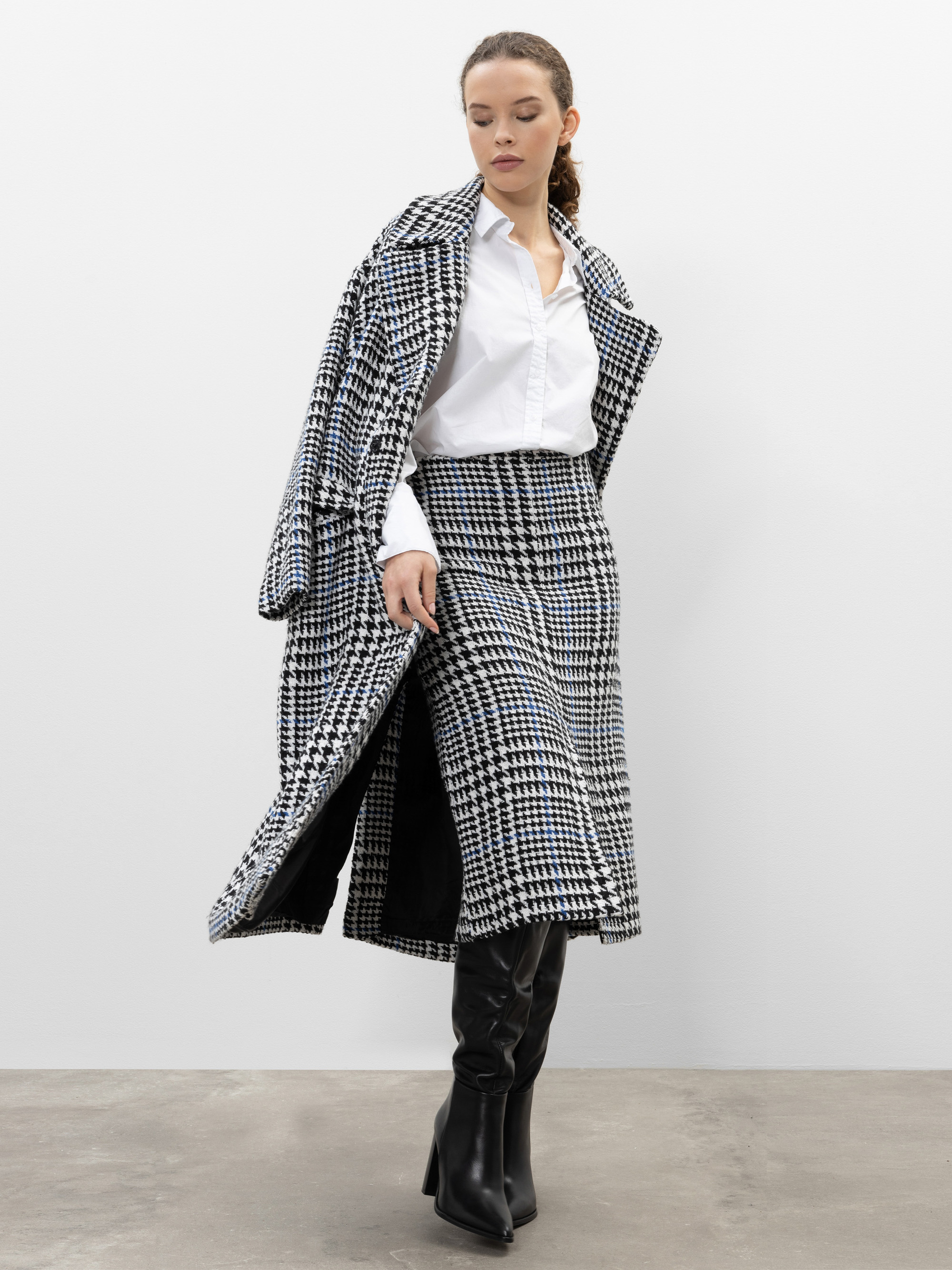Eine Frau trägt einen schwarz-weißen Mantel mit Hahnentrittmuster über den Schultern, einen passenden Rock, ein weißes Hemd zum Knöpfen und schwarze kniehohe Stiefel vor einem schlichten hellen Hintergrund.