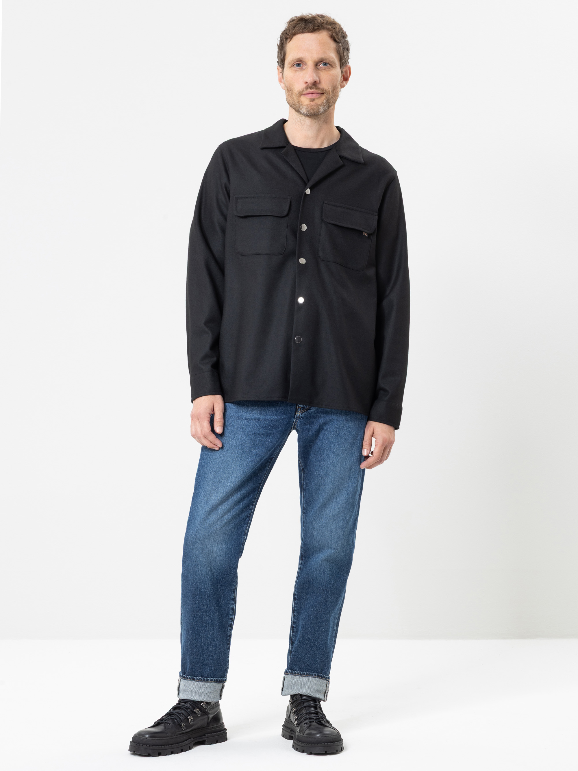 Vor einem weißen Hintergrund steht ein Mann, der ein schwarzes Hemd mit Knöpfen und Brusttaschen, eine blaue Jeans mit umgeschlagenen Beinen und schwarze Schuhe trägt.