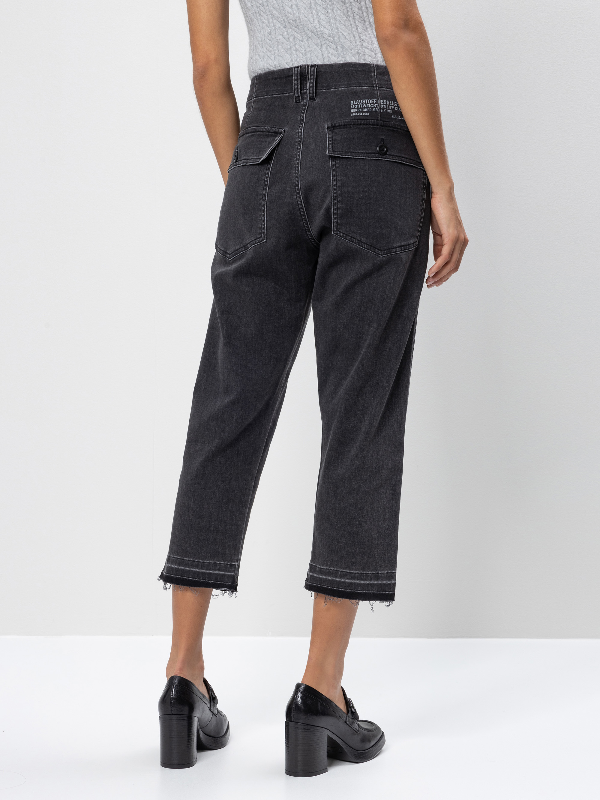 Eine Person, die schwarze Jeans mit ausgefransten Säumen, schwarze Schuhe mit Absatz und ein hellgraues, geripptes, ärmelloses Oberteil trägt, steht mit dem Rücken zur Kamera vor einem weißen Hintergrund.