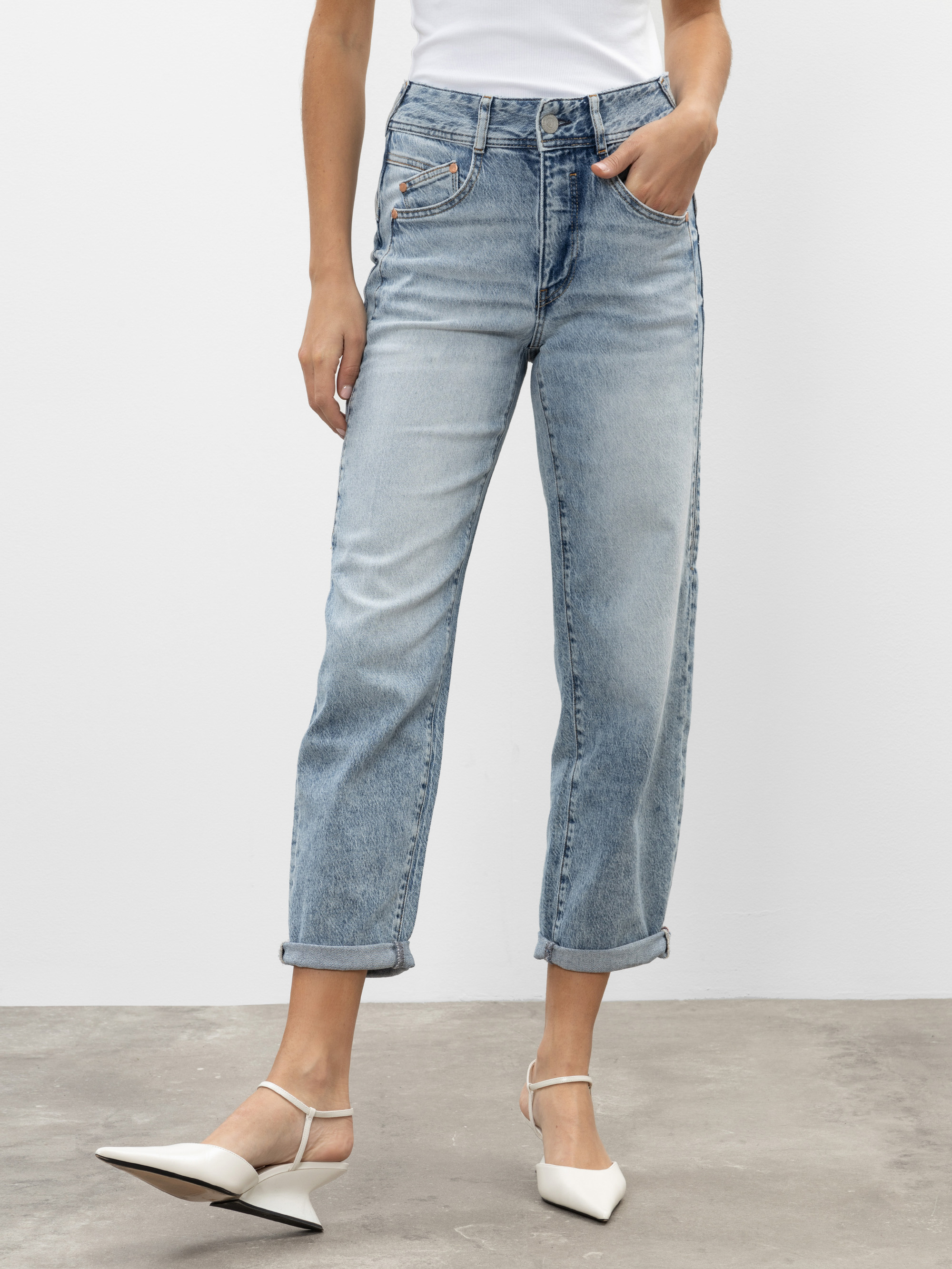 Eine Person, die eine hellblaue Jeans mit hoher Taille und geradem Bein mit aufgerollten Manschetten, ein weißes ärmelloses Oberteil und weiße spitze Absätze trägt, steht vor einem schlichten hellen Hintergrund.