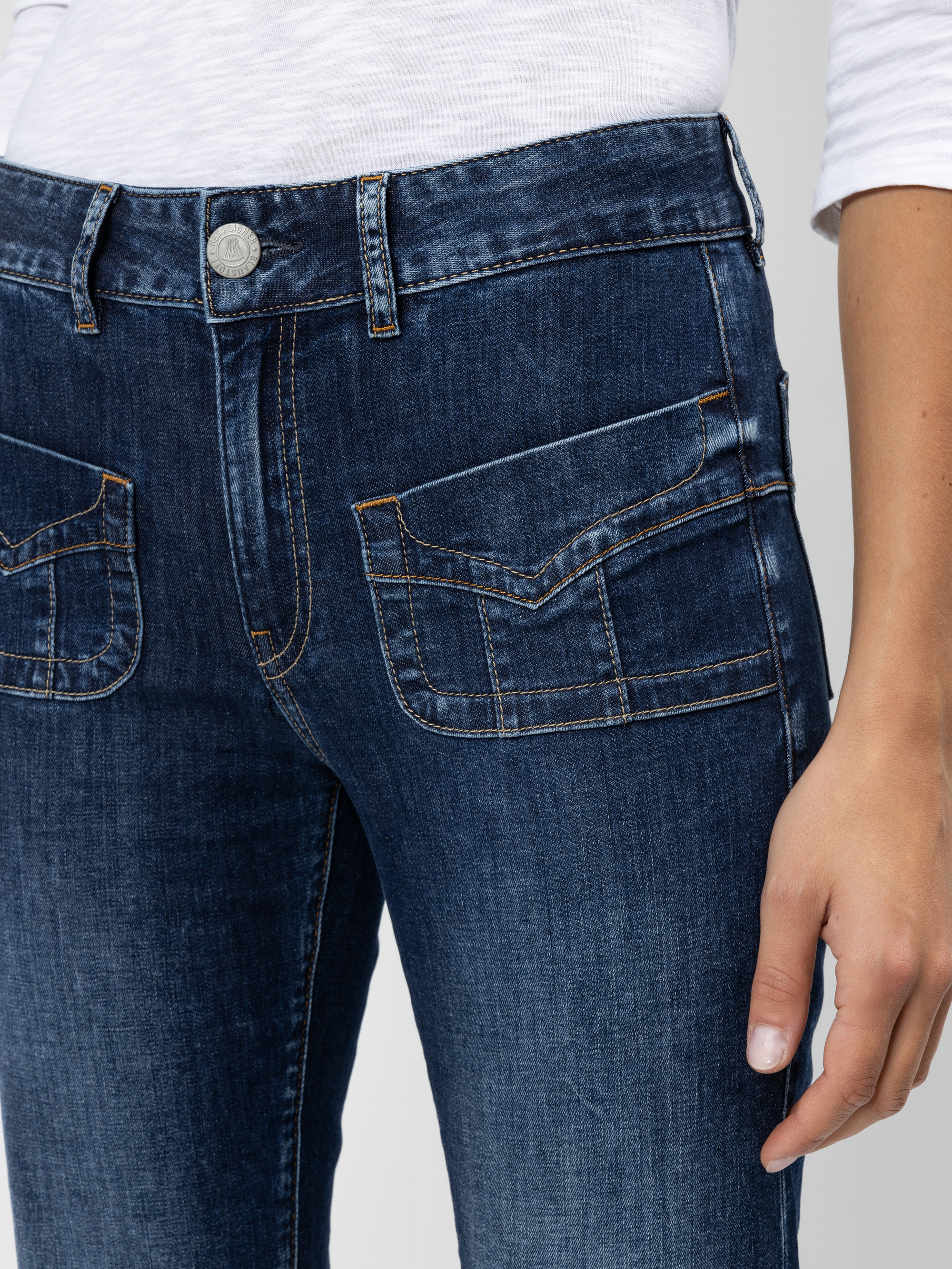 Eine Person, die eine dunkelblaue Jeans mit Klappentaschen und ein weißes langärmeliges Oberteil trägt; die rechte Hand liegt an der Seite.