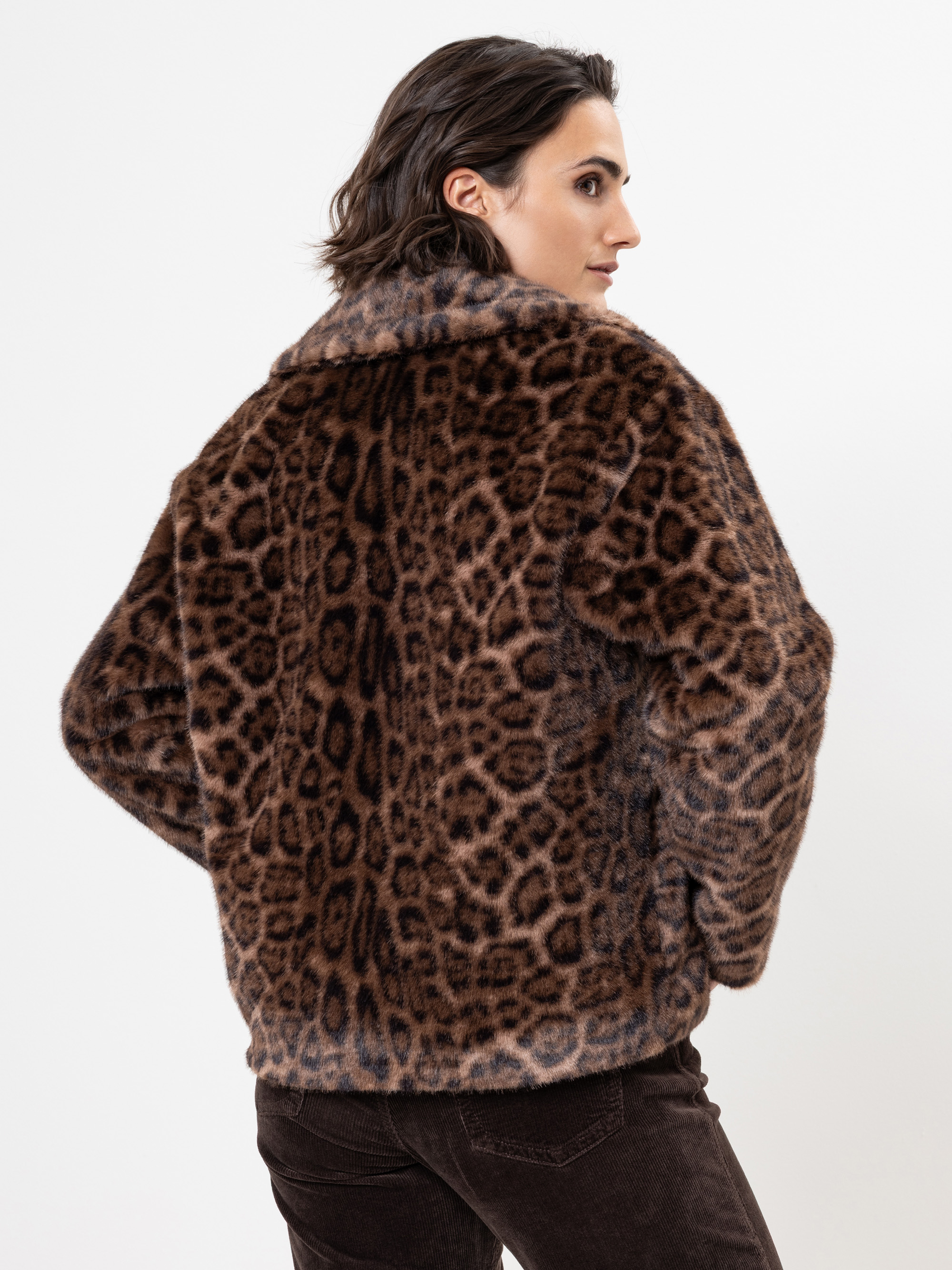 Eine Person mit kurzen dunklen Haaren wird von hinten gezeigt, die eine Jacke aus Kunstpelz mit braunem und schwarzem Leopardenmuster und eine dunkle Hose trägt und vor einem schlichten weißen Hintergrund steht.