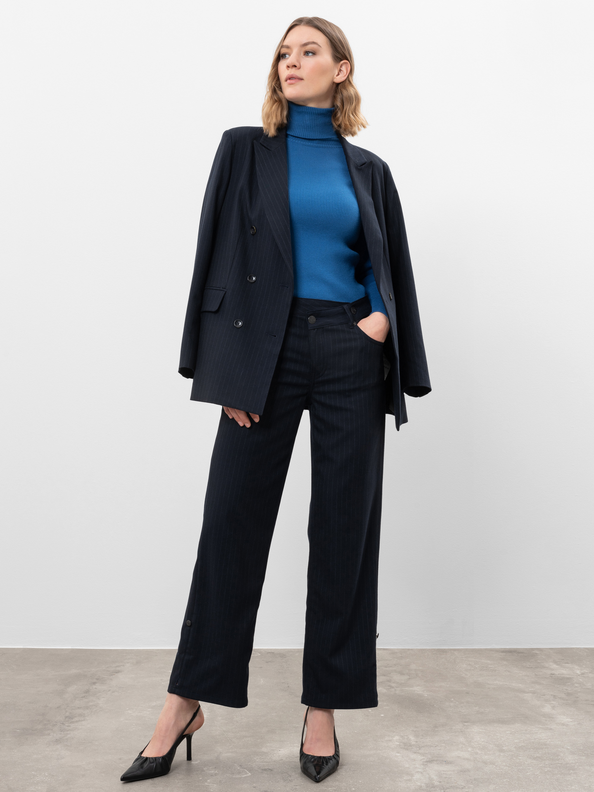 Eine Frau steht selbstbewusst in einem marineblauen Anzug, den Blazer über die Schultern drapiert, mit einem blauen Rollkragenpullover, schwarzen Stöckelschuhen und einer Hose mit geradem Bein vor einem schlichten weißen Hintergrund.