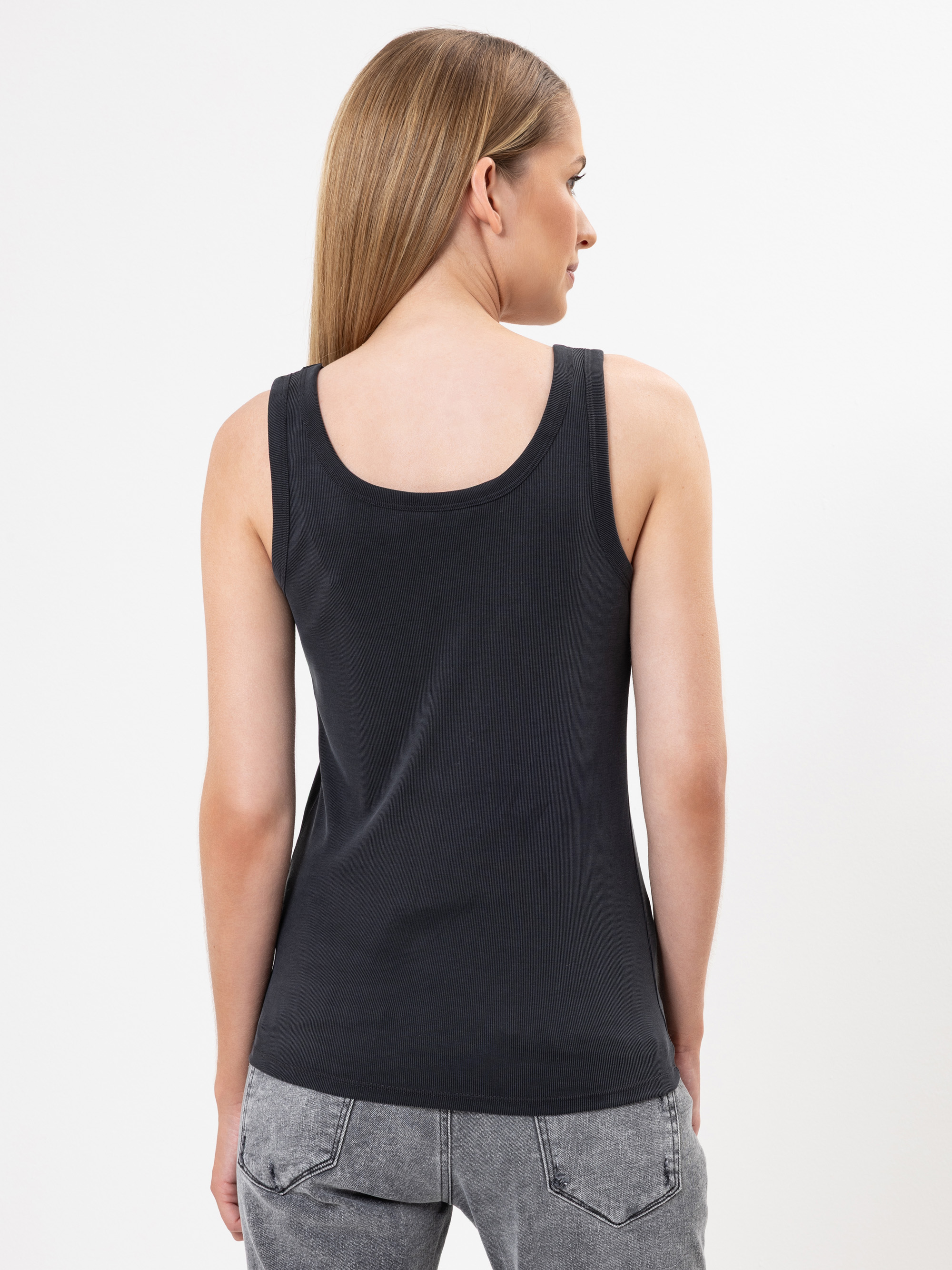 Eine Frau mit glattem, hellbraunem Haar blickt von der Kamera weg und trägt ein ärmelloses schwarzes Oberteil und graue Jeans. Sie steht vor einem schlichten weißen Hintergrund.