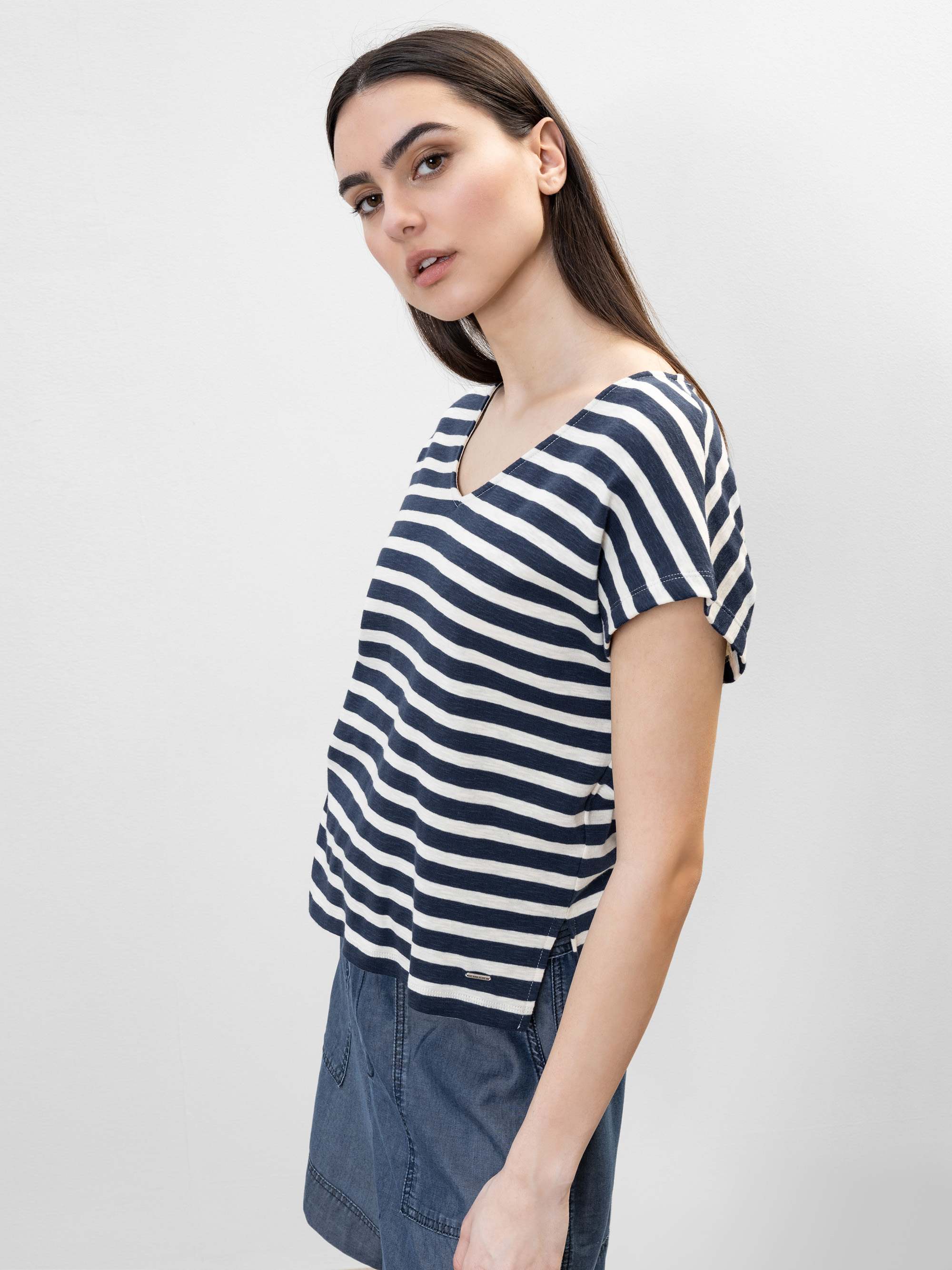 Eine Frau mit langen dunklen Haaren trägt ein marineblau und weiß gestreiftes T-Shirt mit V-Ausschnitt und einen blauen Rock. Sie steht seitlich und blickt leicht über die Schulter vor einem schlichten hellen Hintergrund.