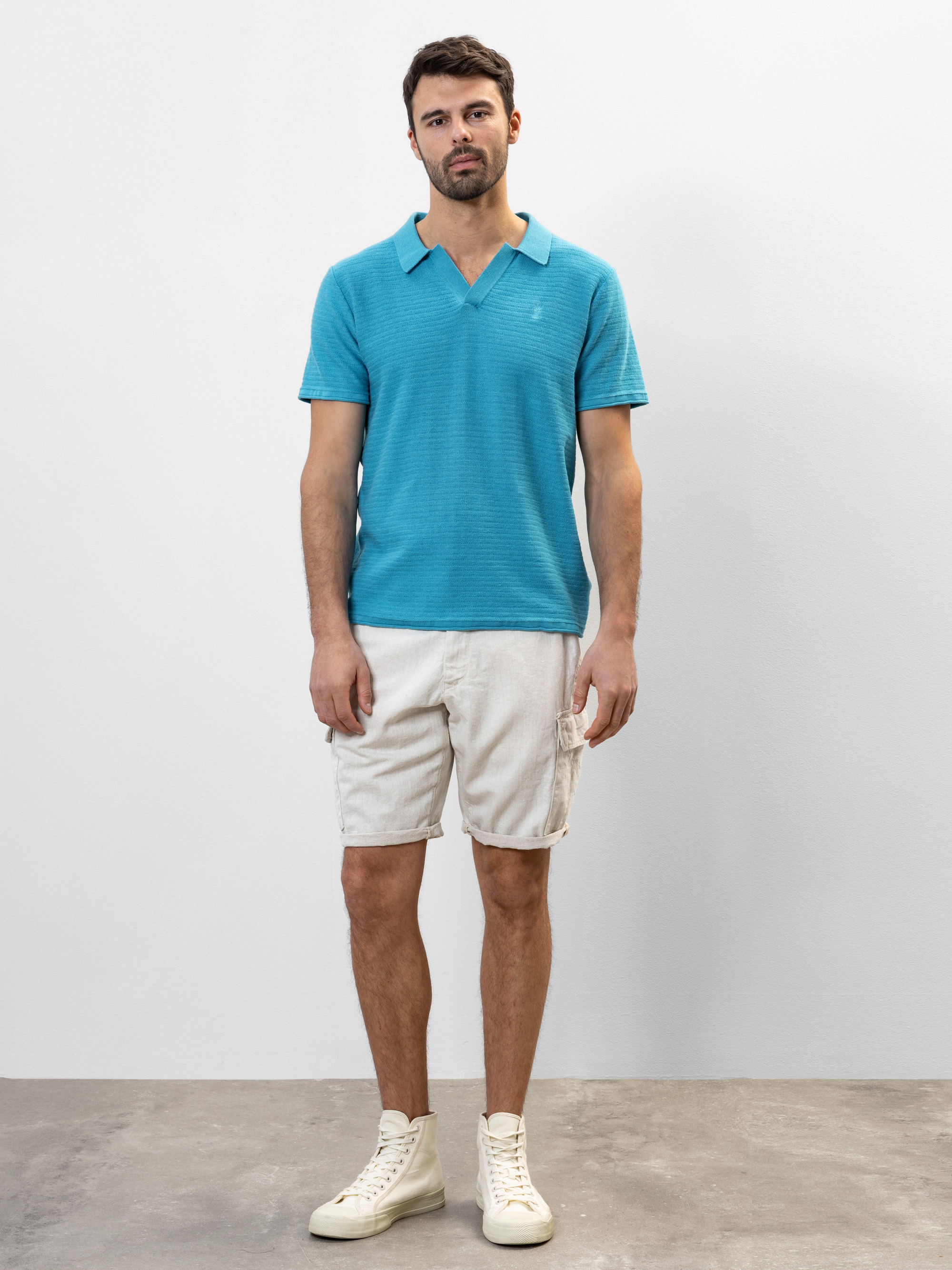Ein Mann steht vor einer schlichten weißen Wand und trägt ein türkisfarbenes, kurzärmeliges Polohemd, beigefarbene Cargoshorts und weiße Turnschuhe mit hohen Absätzen. Er blickt mit neutralem Gesichtsausdruck und entspannter Körperhaltung nach vorn
