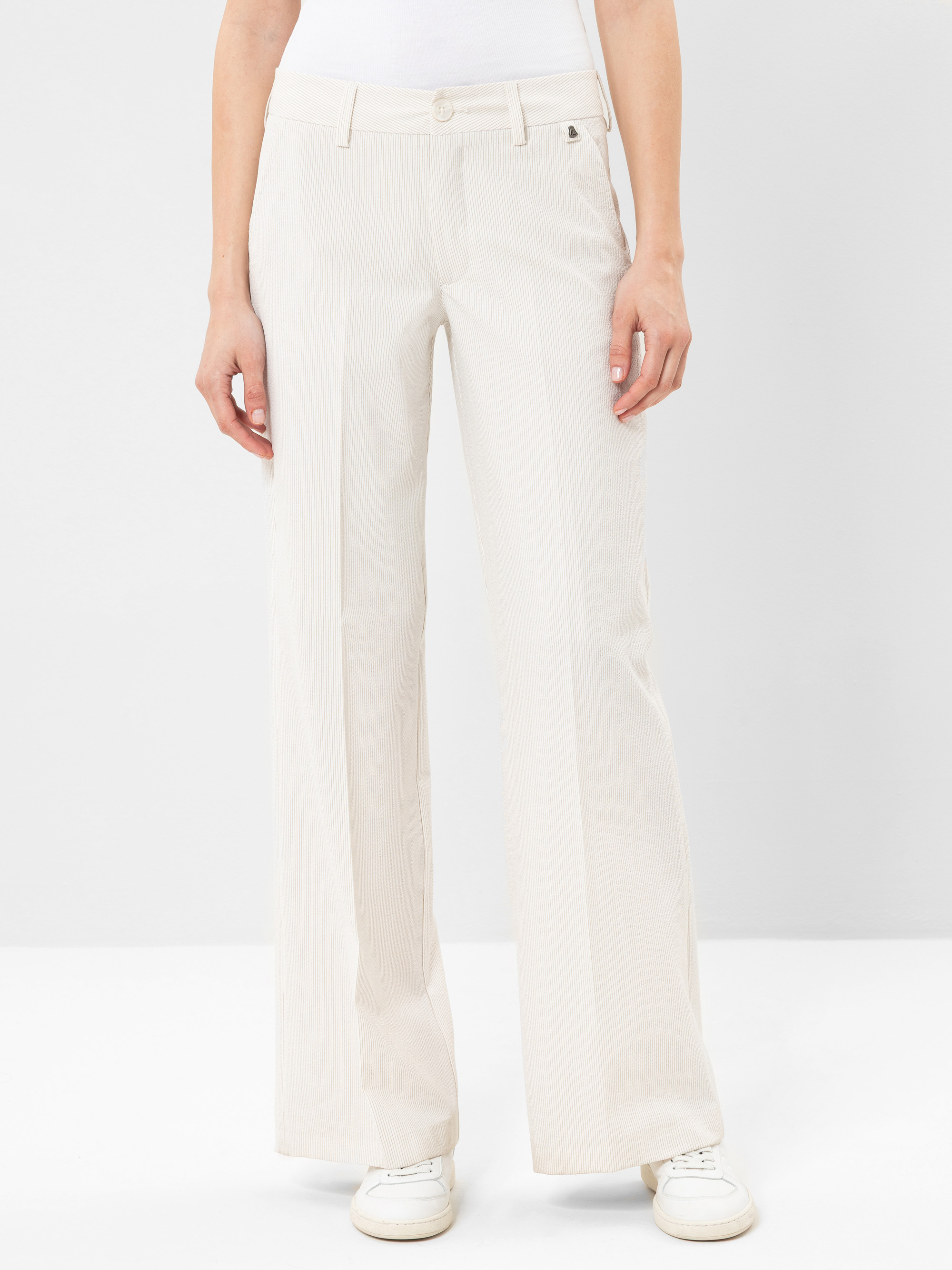 Eine Person, die eine hellbeige Hose mit hoher Taille und weitem Bein mit dezentem Nadelstreifenmuster, weiße Turnschuhe und ein weißes Oberteil trägt, steht vor einem schlichten hellen Hintergrund.