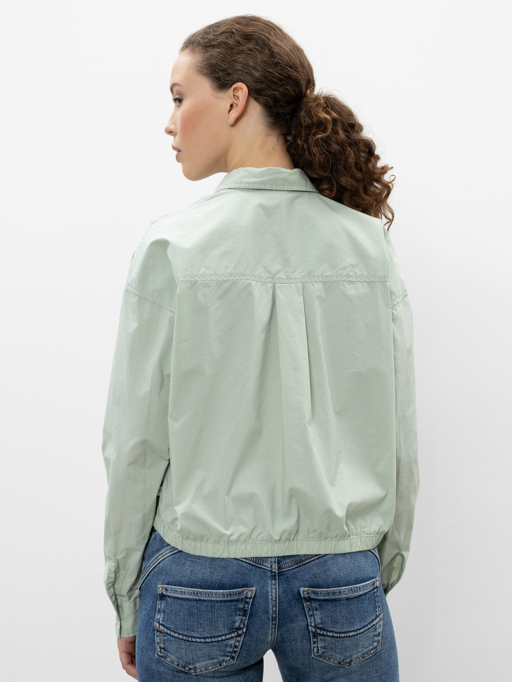 Eine Frau mit lockigem, zurückgebundenem braunem Haar trägt eine blassgrüne, langärmelige Jacke und blaue Jeans. Sie steht mit dem Rücken zur Kamera vor einem schlichten weißen Hintergrund.