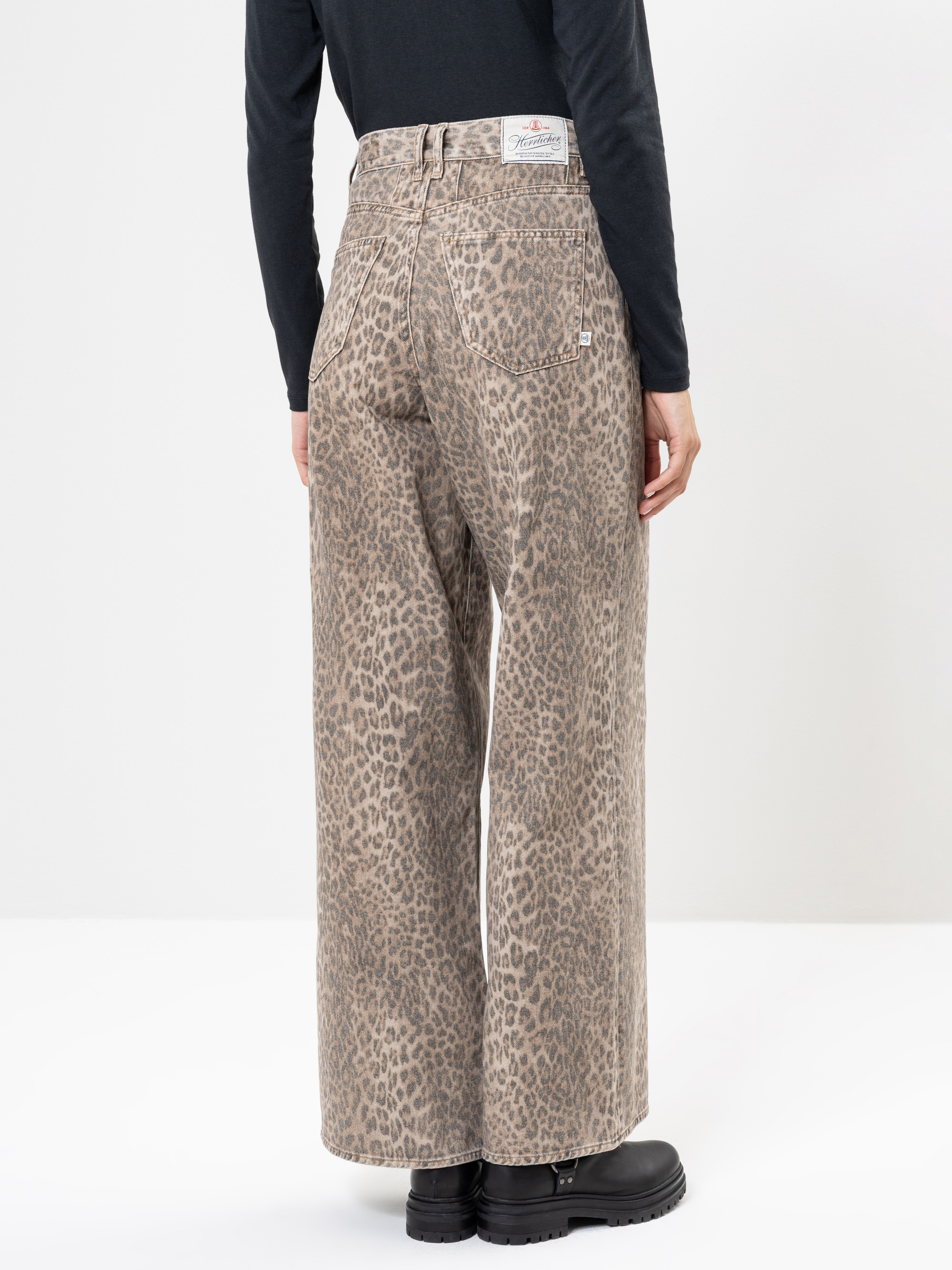 Eine Person, die eine hellbraune Jeans mit weitem Bein und hoher Taille mit Leopardenmuster, ein schwarzes langärmeliges Oberteil und schwarze Stiefel trägt, steht mit dem Gesicht nach unten vor einem schlichten weißen Hintergrund.