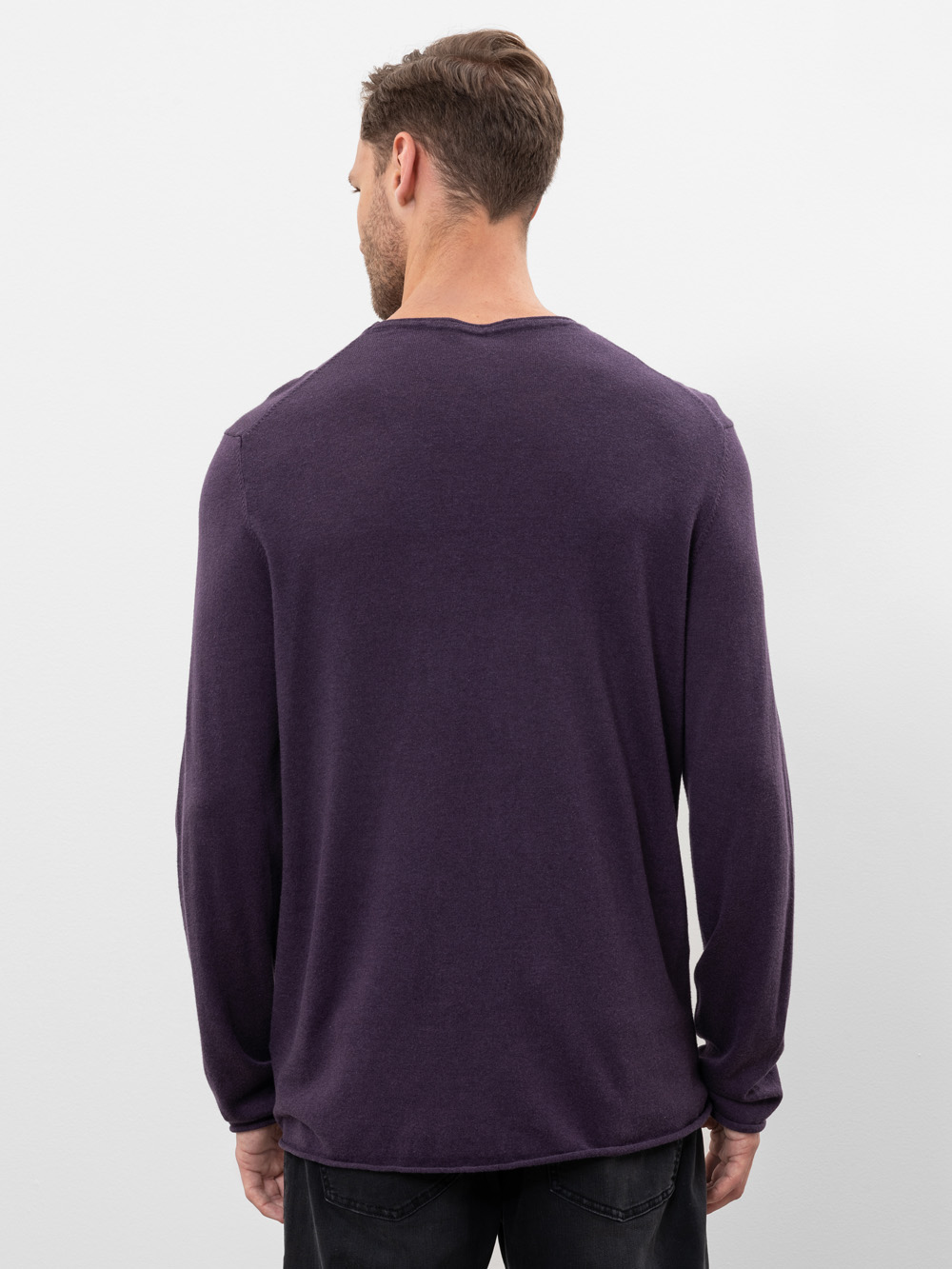 Ein Mann mit kurzen braunen Haaren steht mit dem Rücken zur Kamera, trägt ein langärmeliges, dunkelviolettes Hemd und eine schwarze Hose vor einem schlichten weißen Hintergrund.