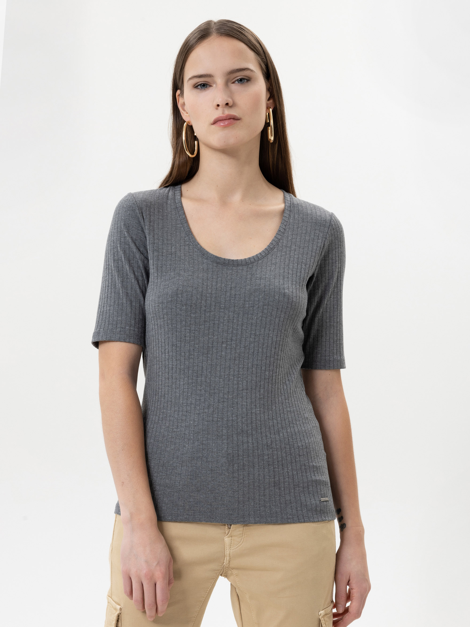 Eine Frau mit glattem braunem Haar, die goldene Ohrringe, ein geripptes graues kurzärmeliges Oberteil und eine beigefarbene Hose trägt, steht vor einem schlichten weißen Hintergrund und schaut direkt in die Kamera.